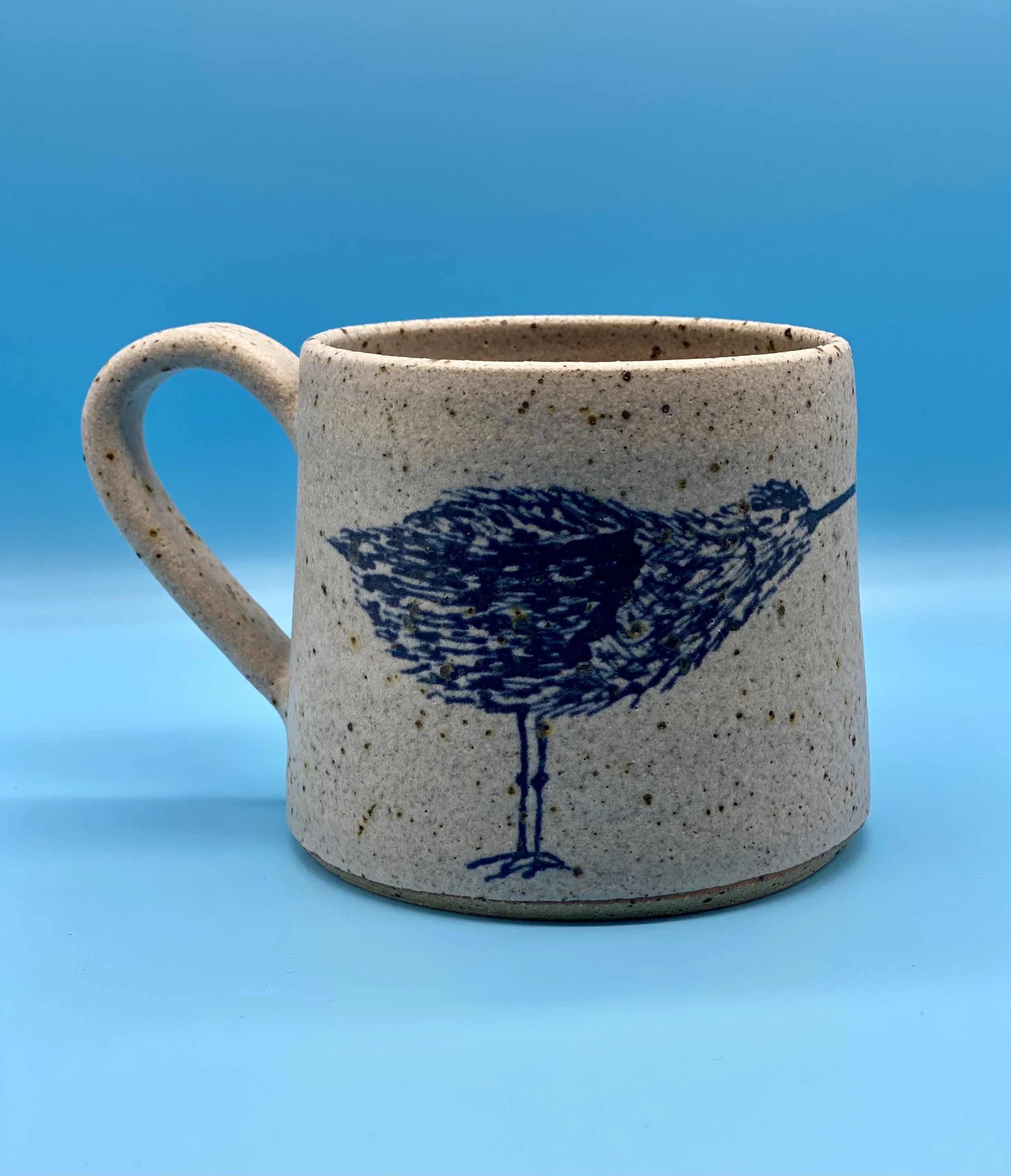 A speckled ceramic mug with a blue bird design, placed on a light blue surface against a bright blue background.