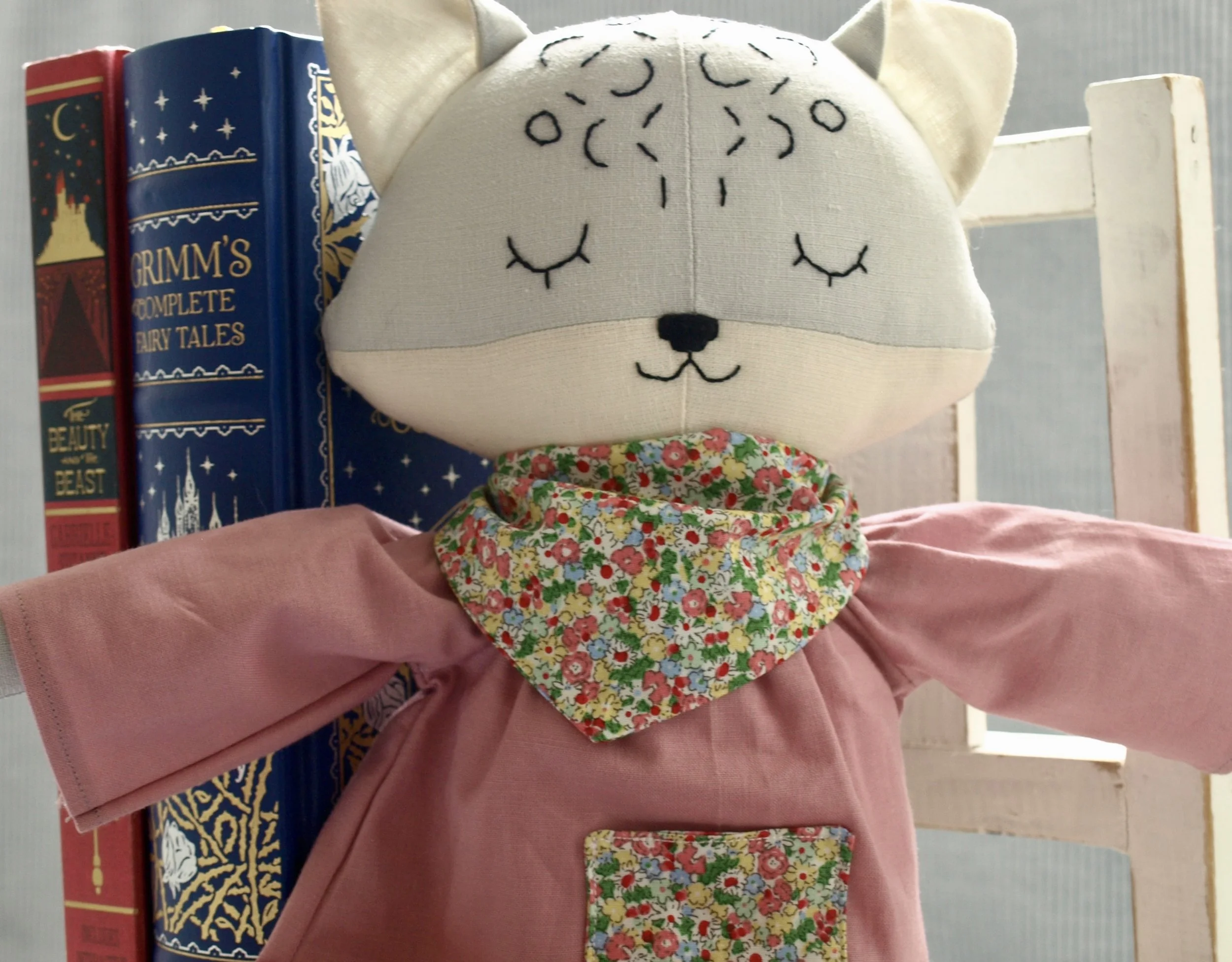 A plush stuffed animal with closed eyes and embroidered black details, wearing a colourful floral bandana around its neck, with a pink dress, placed on a wooden shelf next to colourful books.