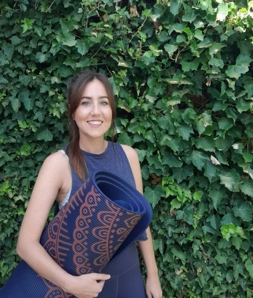 A young woman holding a rolled yoga mat standing in front of a leafy green hedge.