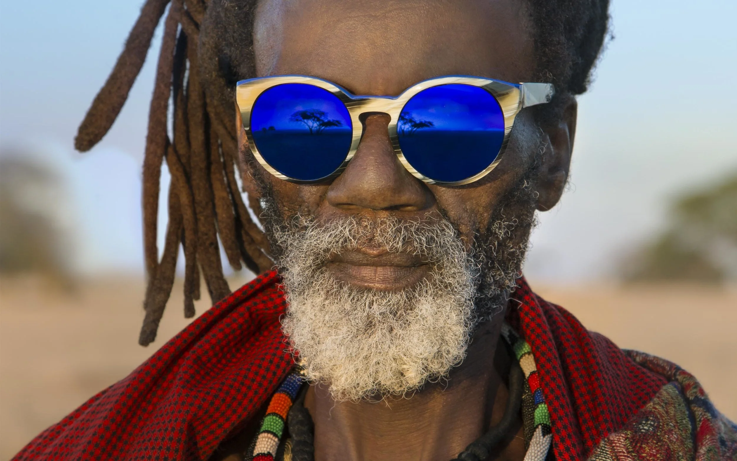 Stylishly dressed black man with white beard and dreadlocks wearing sunglasses that are reflecting a vast landscape with a couple of trees.