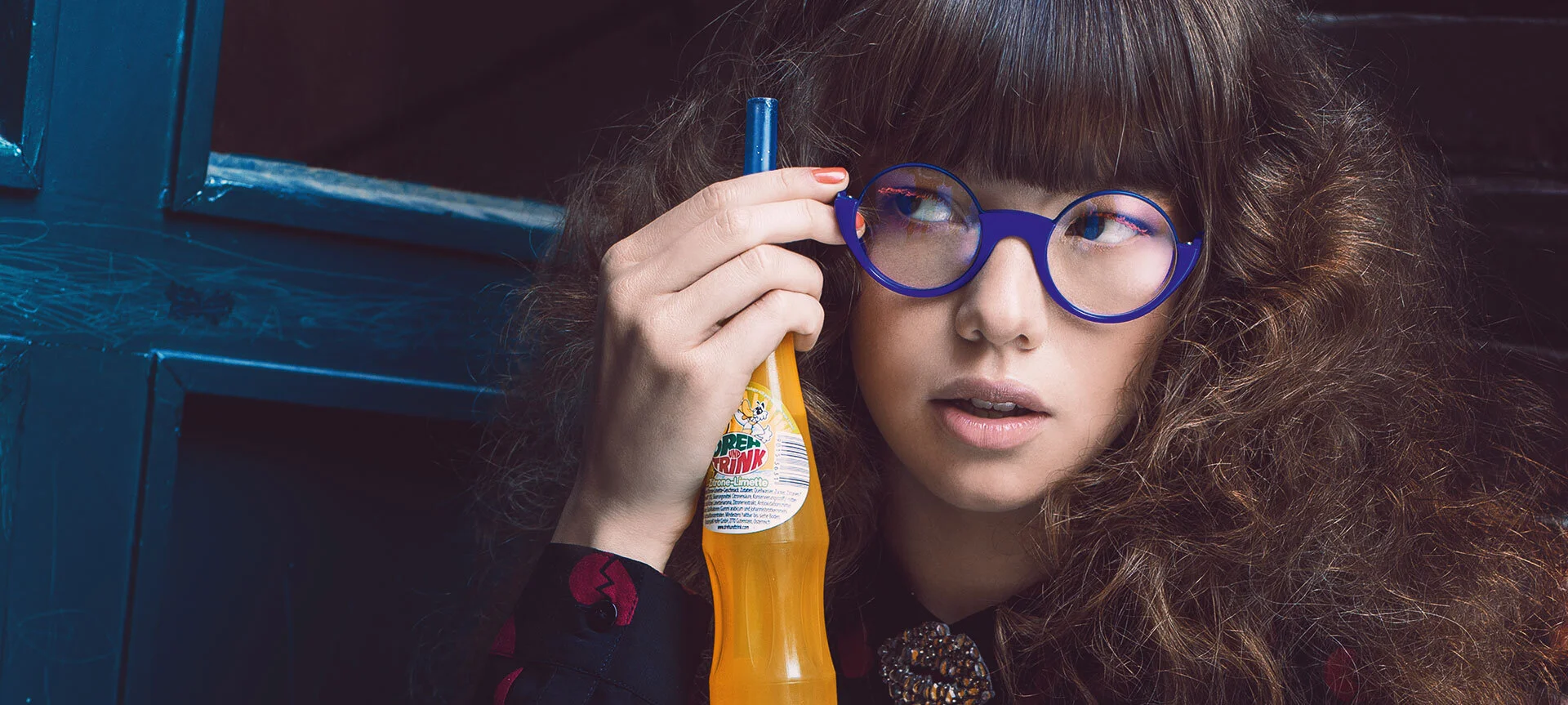 Stylish young woman in blue eyeglasses with curly hair and short bangs next to dark blue door holding an orange soda bottle with straw
