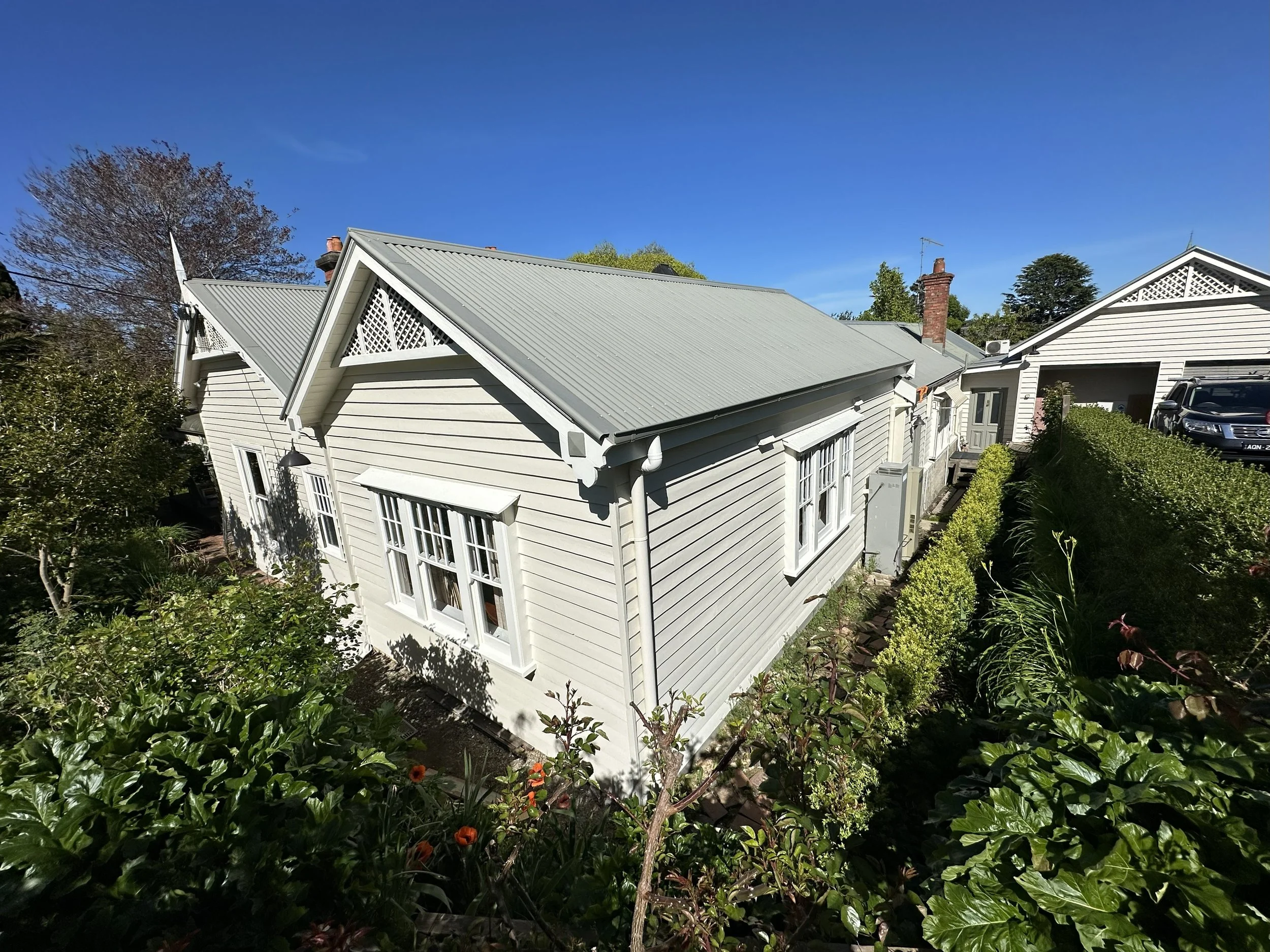 View of a white house with a gray metal roof, surrounded by green shrubs and trees, under a clear blue sky.