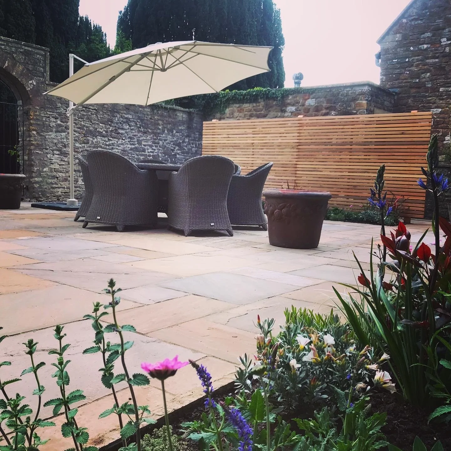 Outdoor patio with a dining table, four wicker chairs, a large white umbrella, potted plants, and a wooden fence with stone walls.