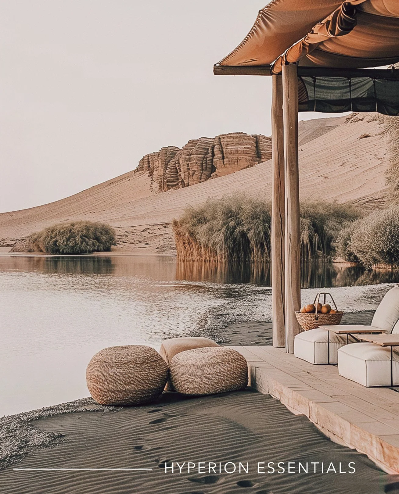 A desert landscape with sand dunes, large rocks, and sparse desert vegetation near a body of water. There is a wooden structure with outdoor seating, including cushioned chairs and woven ottomans, beside the water, under a canopy. Mountains or large rock formations are in the background.
