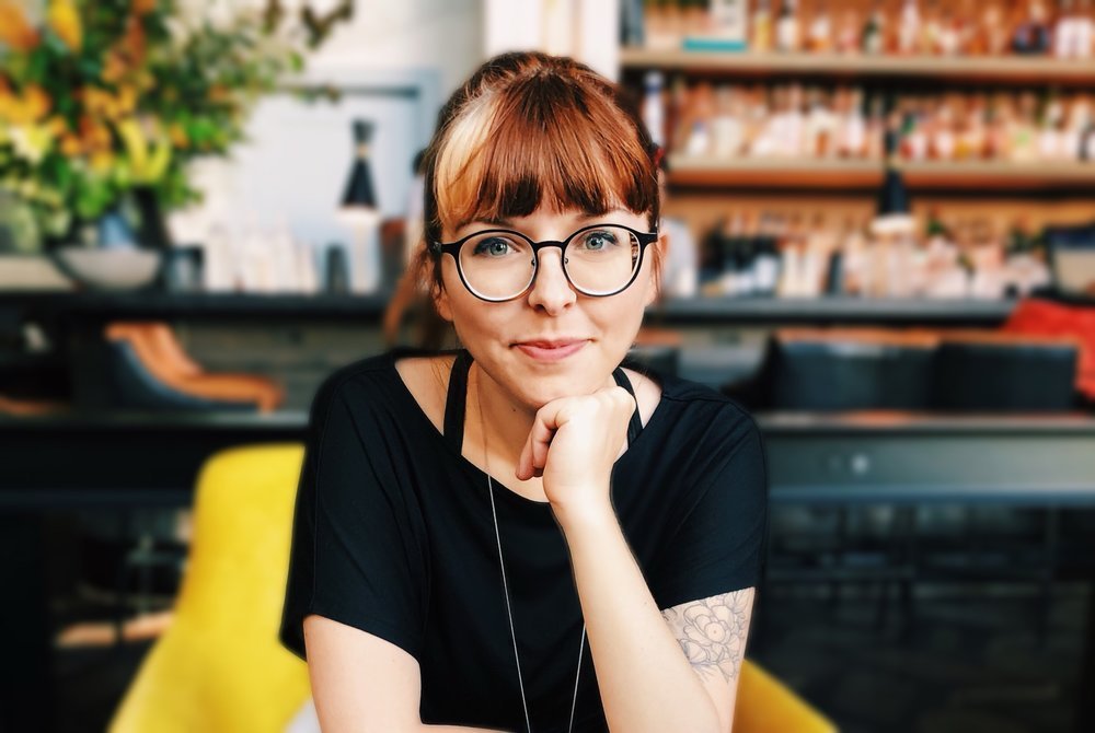 A woman with red hair, glasses, and a black shirt sitting in a cafe or bookstore, resting her chin on her hand, with a blurred background of bookshelves and plants.