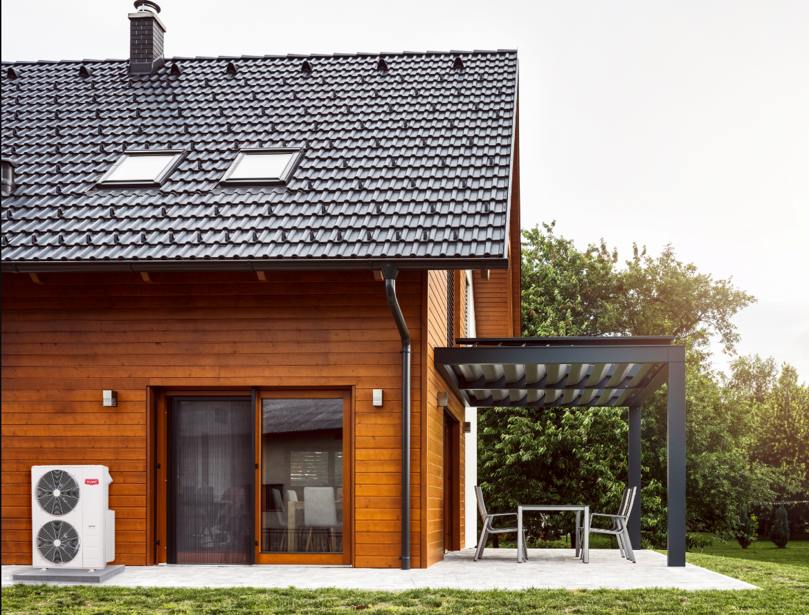 A modern house with a brown wooden exterior, black roof tiles, solar skylights, a chimney, and an outdoor patio with a table and two chairs, surrounded by green trees and grass.