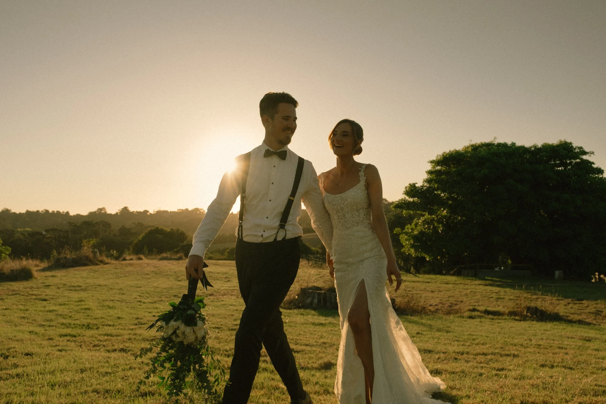 A bride and groom walk together in a field in Auckland, smiling, with the groom holding the bride's bouquet of flowers.