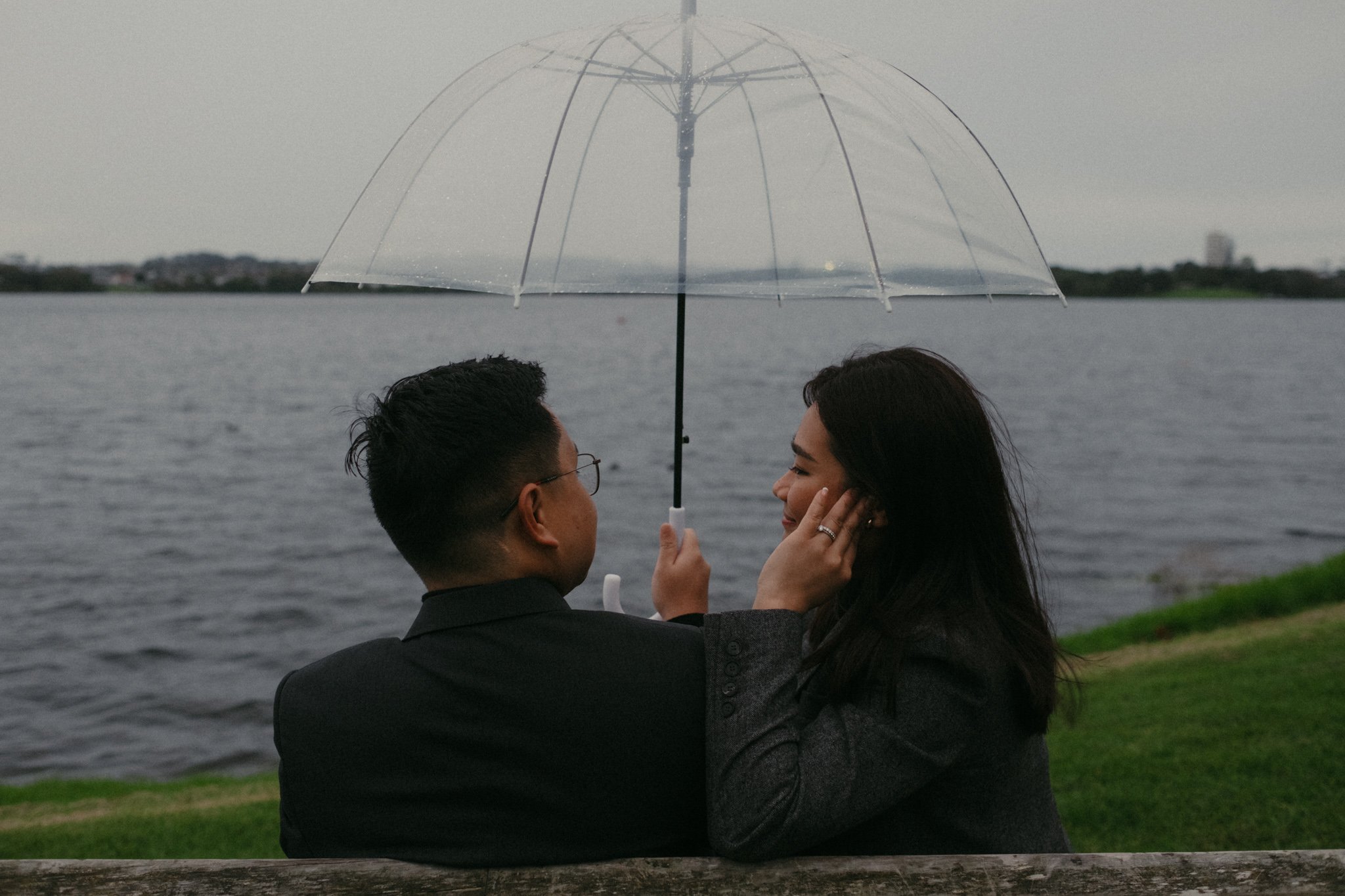 A couple sitting on a bench by the water in Lake Pupuke, Auckland, sharing a transparent umbrella during overcast weather.