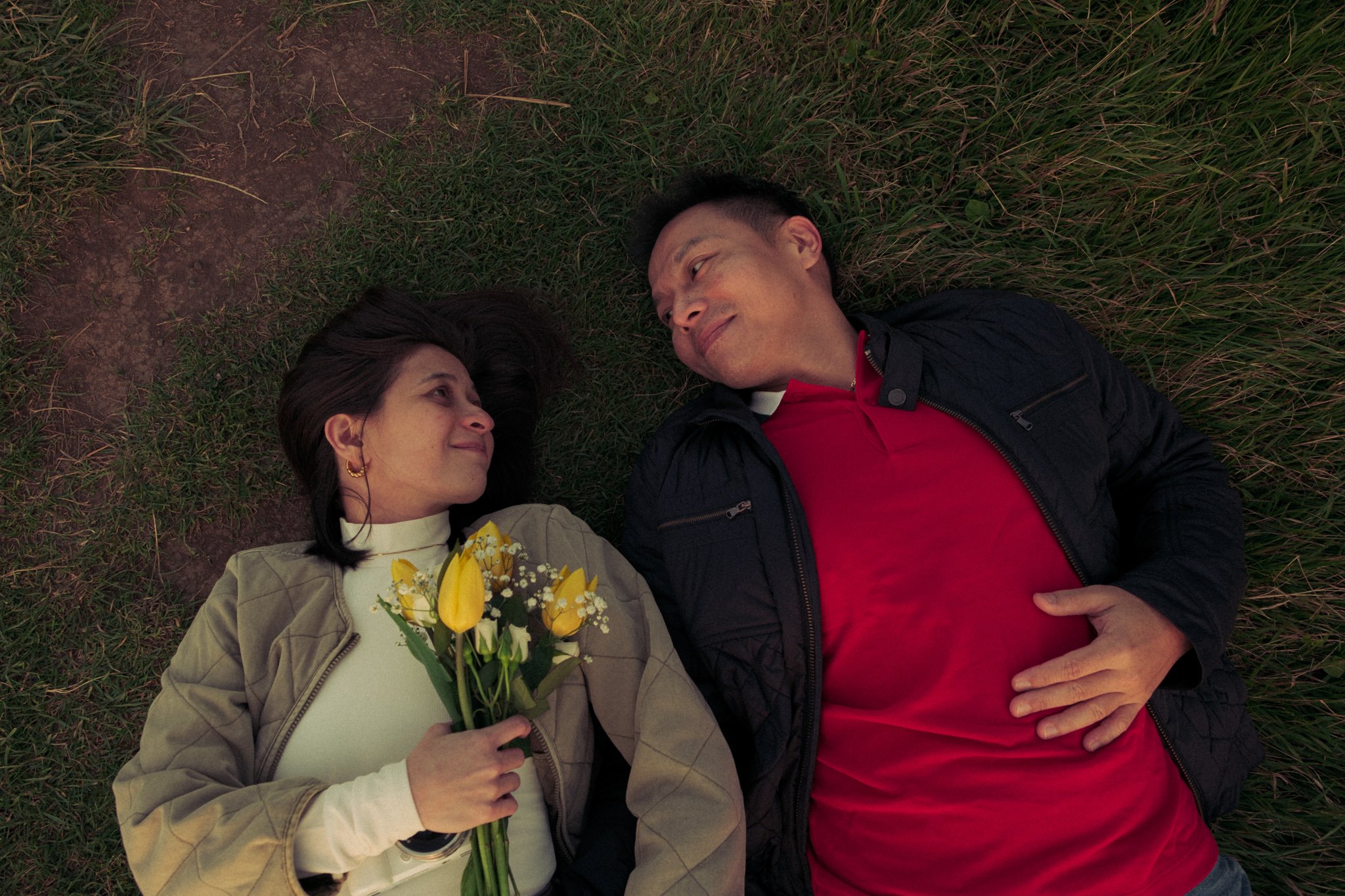 A man and woman lying on grass, looking at each other, with the woman holding a bouquet of yellow flowers.