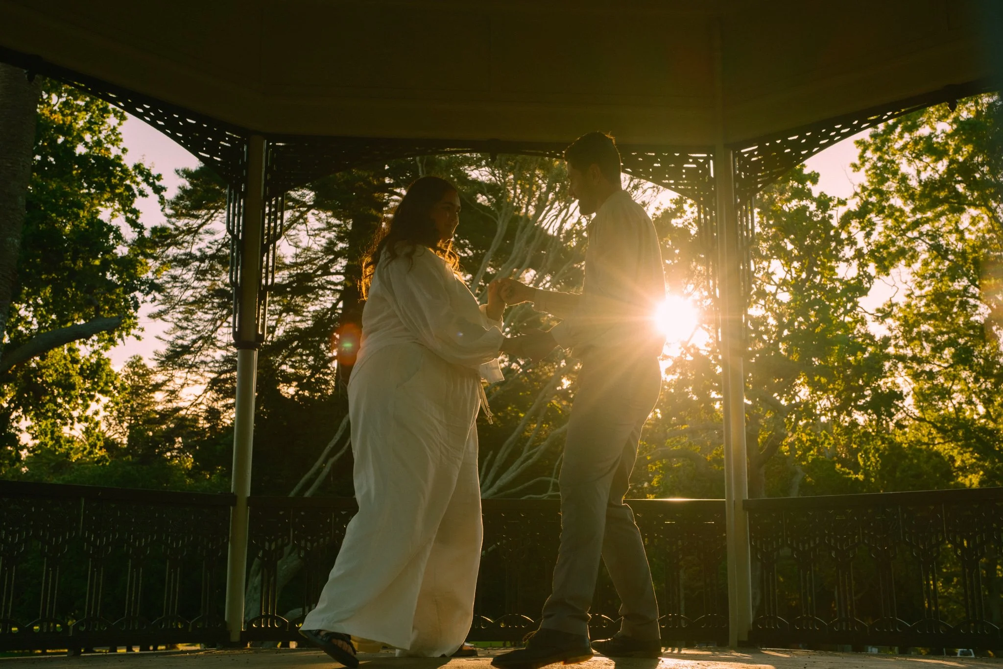 A couple holding hands on a balcony at sunset, surrounded by trees, with the sun shining behind them.
