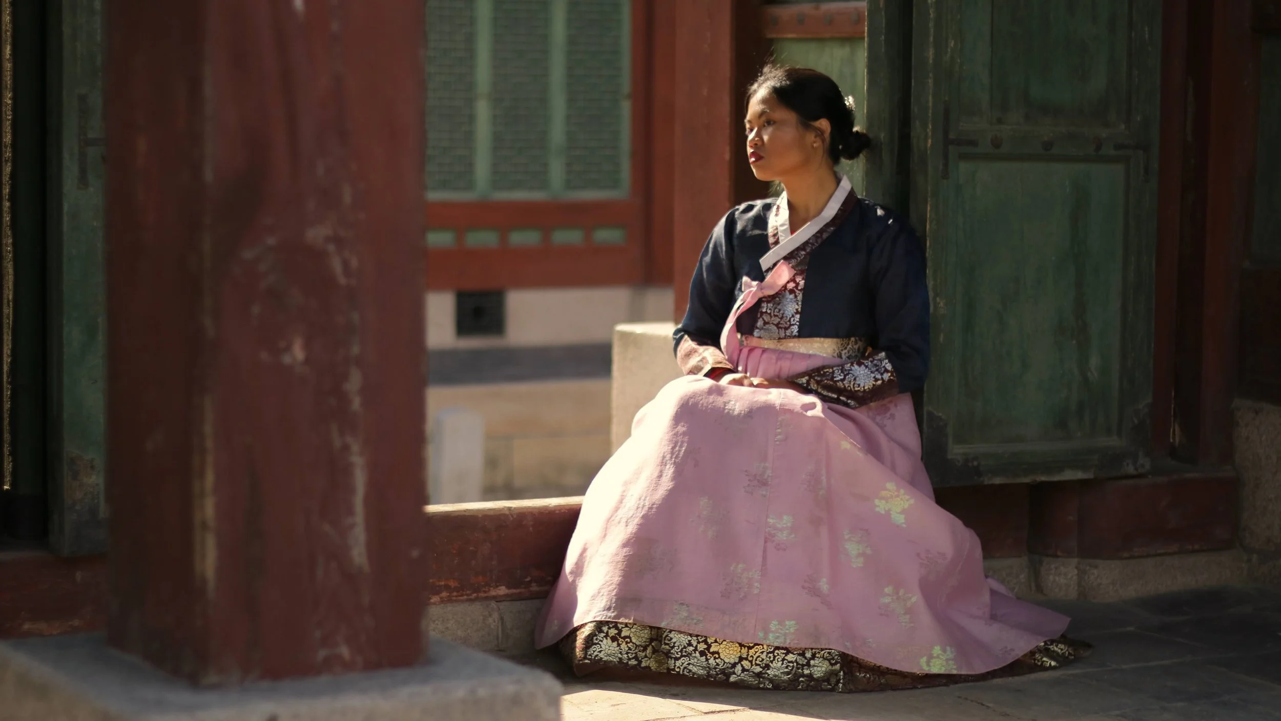A woman wearing traditional Korean hanbok fabric sitting on a stone surface near an open green wooden door at a Korean palace.
