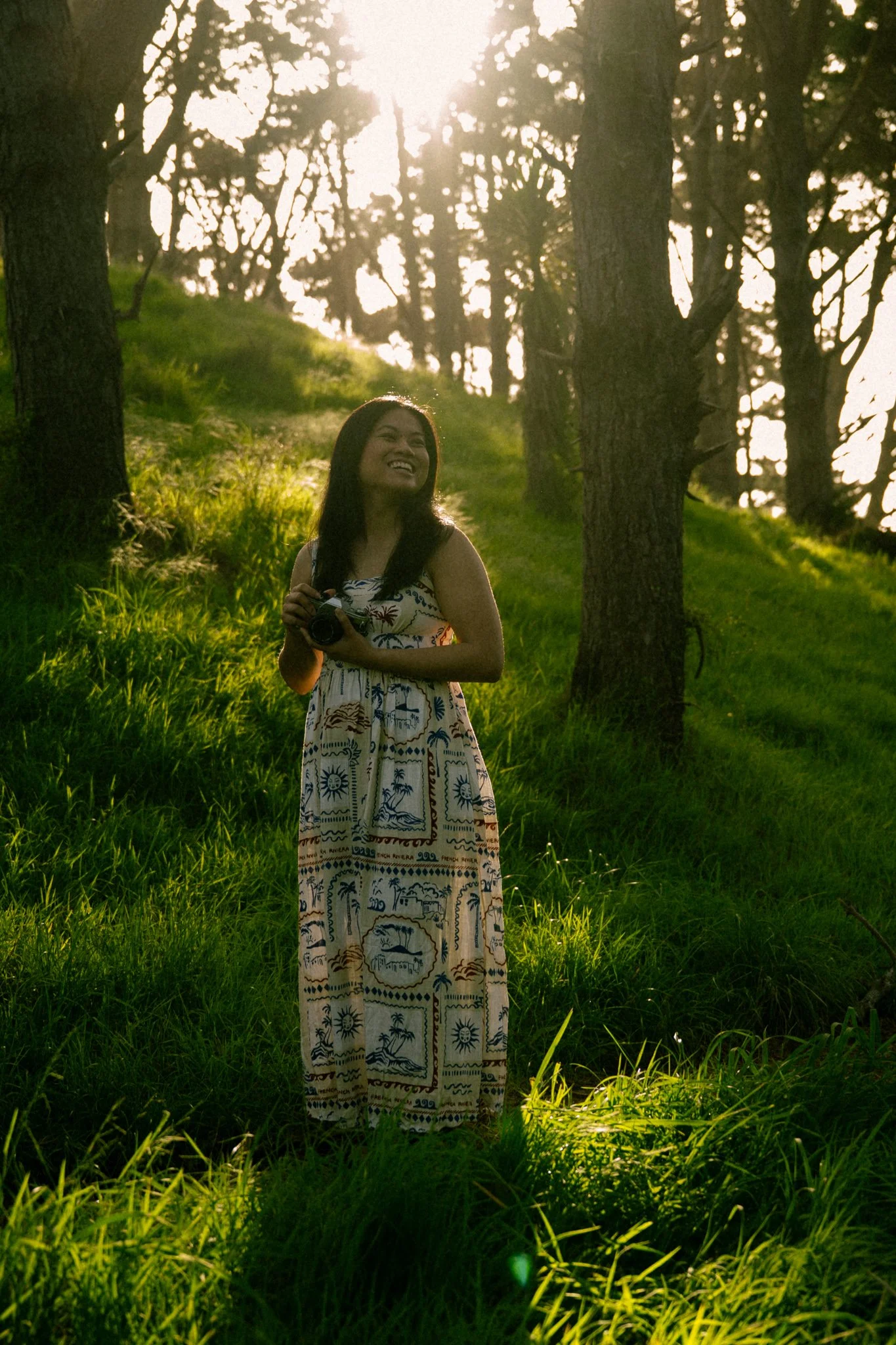 A woman outdoors in a grassy area with trees around her, holding a camera and smiling as sunlight shines through the trees behind her.