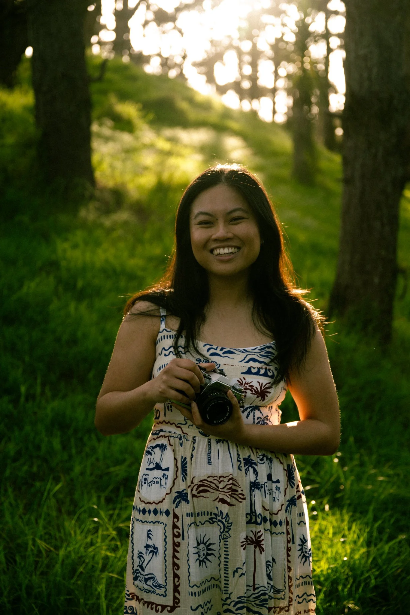 Young woman outdoors in a forest during sunset, holding a camera and smiling.