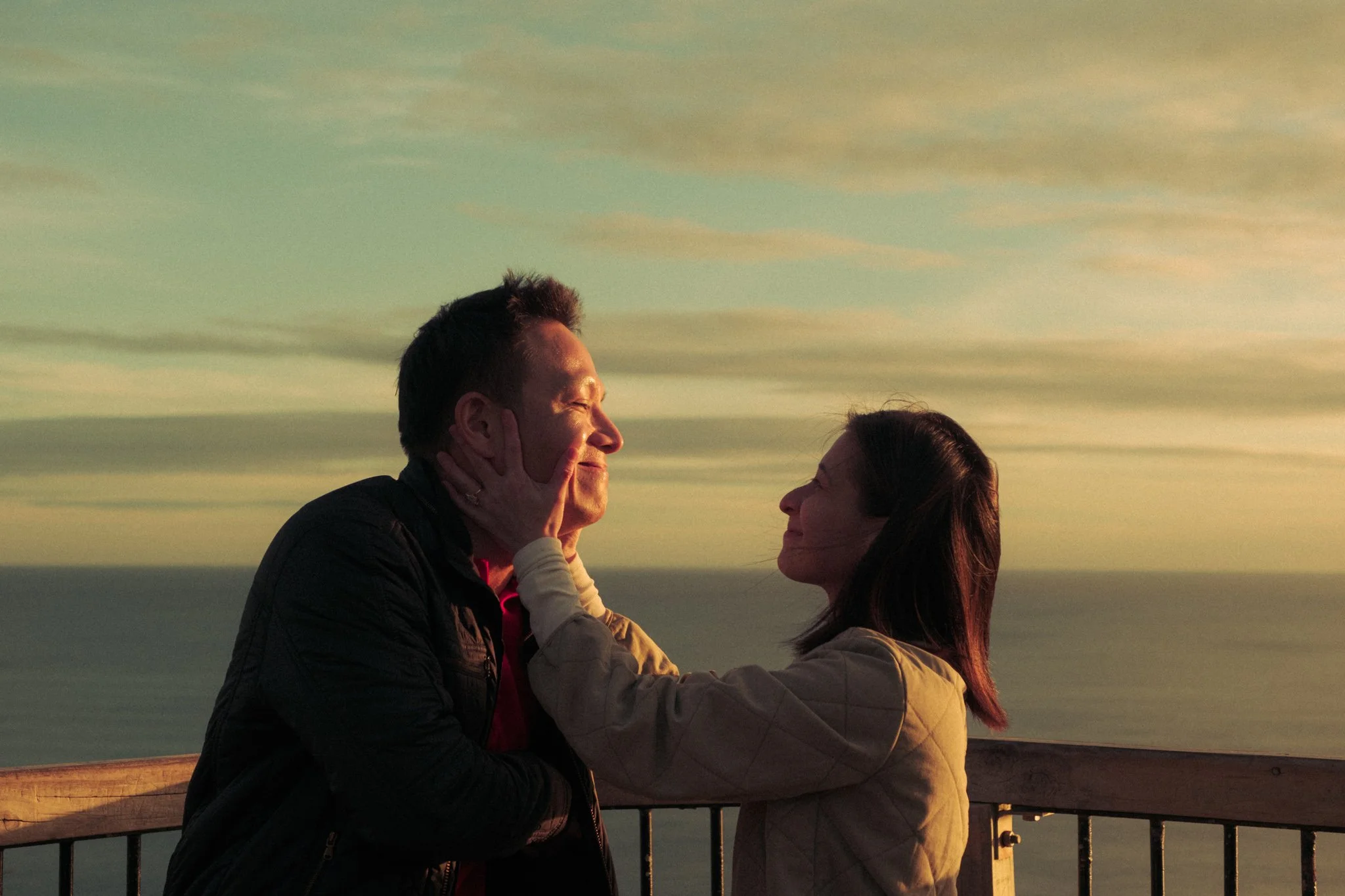 A man and a woman share a romantic moment on a balcony during sunset, with the man touching the woman's face and both smiling at each other, against a backdrop of a colorful sky with clouds.