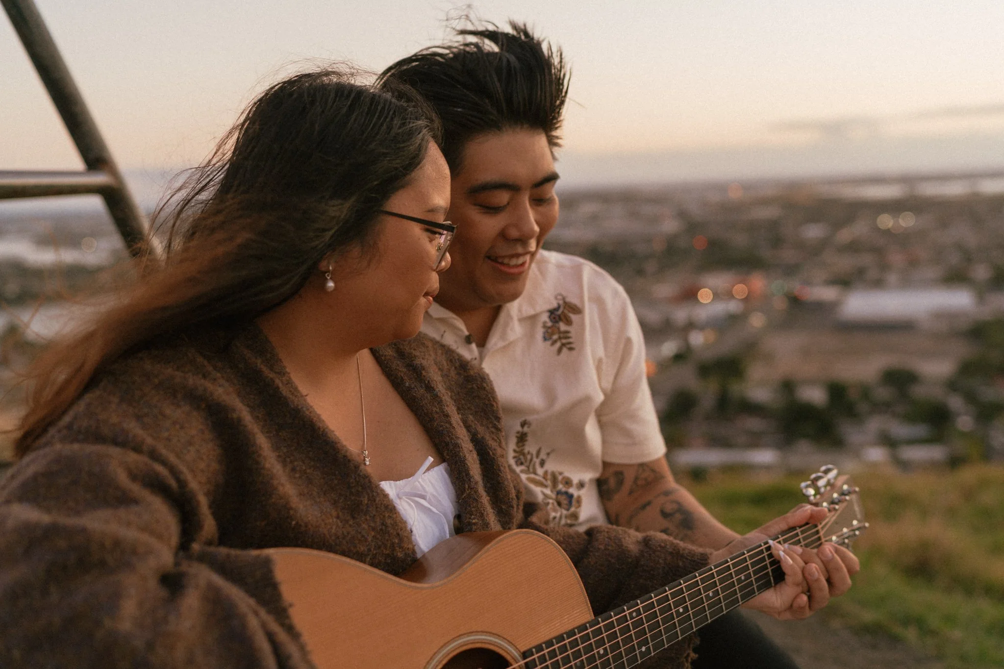 An engaged couple sitting on the Mount Wellington summit at sunset, smiling and playing guitar together with a cityscape in the background.