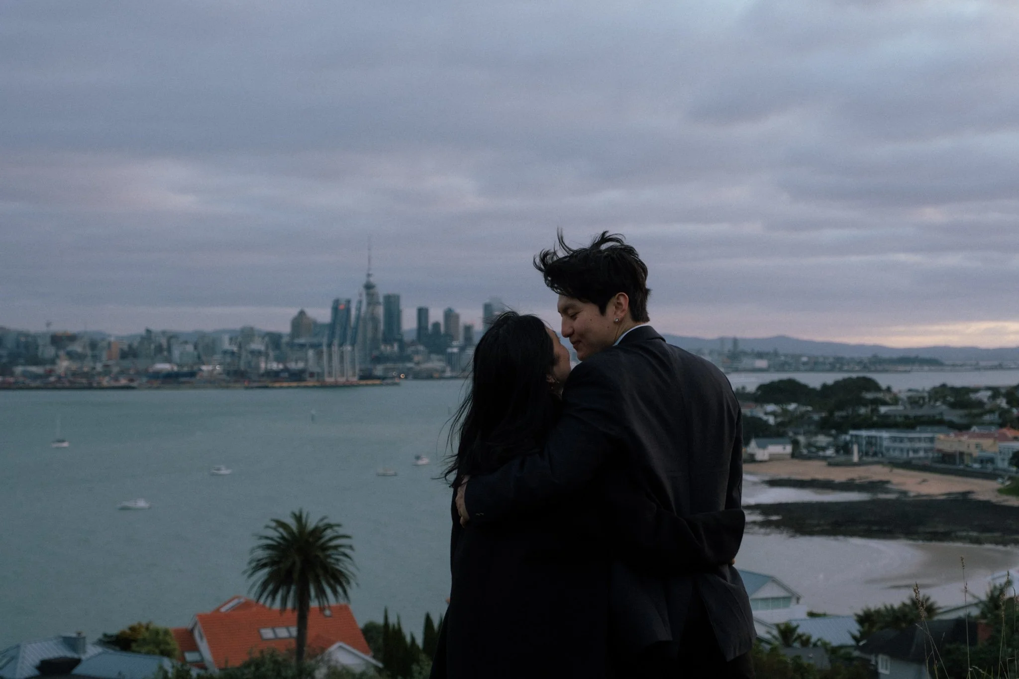 A couple embracing outdoors with a city skyline in the background, overcast sky.