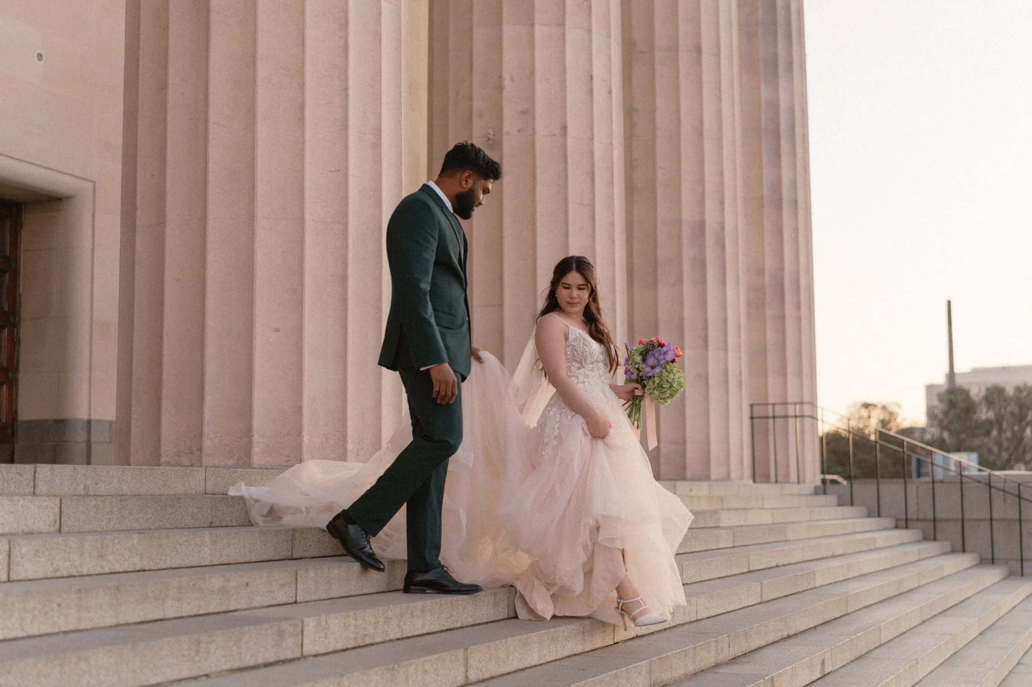 A bride holding a colourful bouquet of flowers steps down the stairs outside Auckland Museum, while her groom assists her.