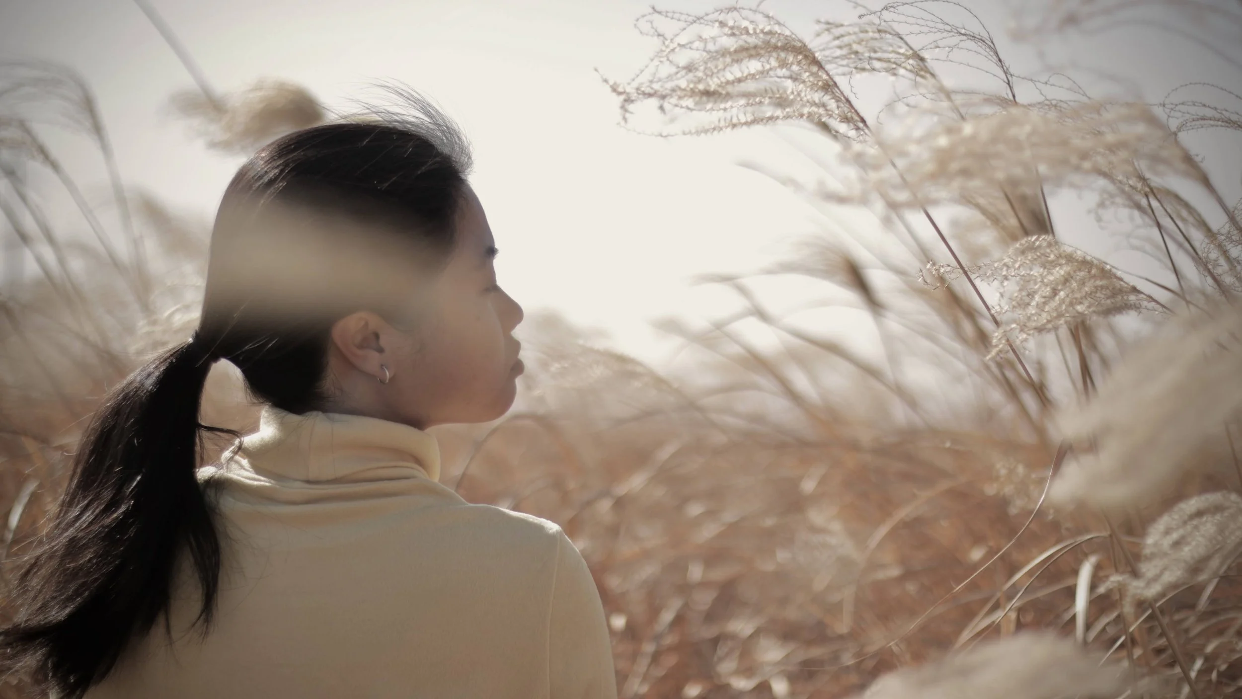 A woman with long dark hair in a ponytail, wearing a light-colored turtleneck, sitting among tall, beige, feathery grasses with her eyes closed, facing to the right, in soft sunlight.
