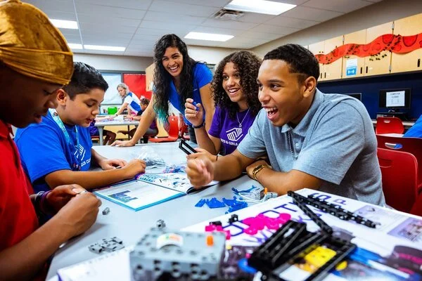A diverse group of students and a teacher laughing and working together at a table in a classroom.