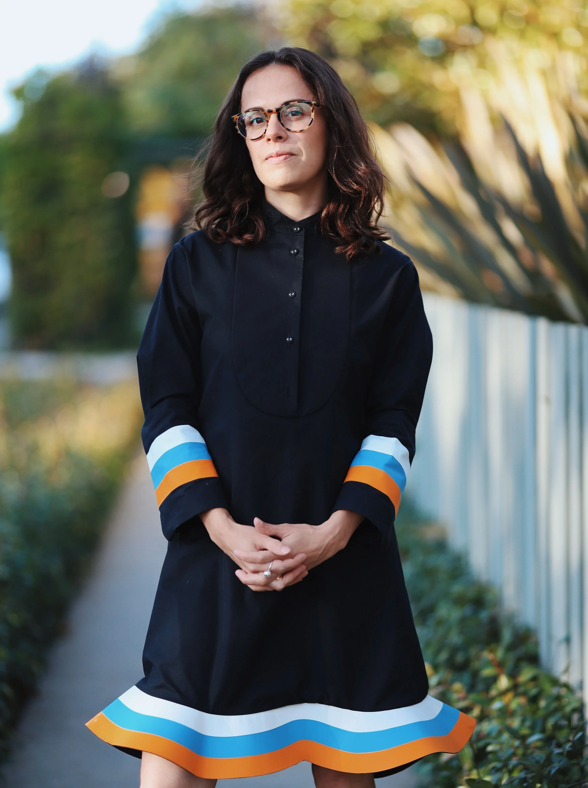A woman with glasses wearing a black dress with colorful striped cuffs and hem, standing outdoors on a pathway with trees and a metal fence in the background.