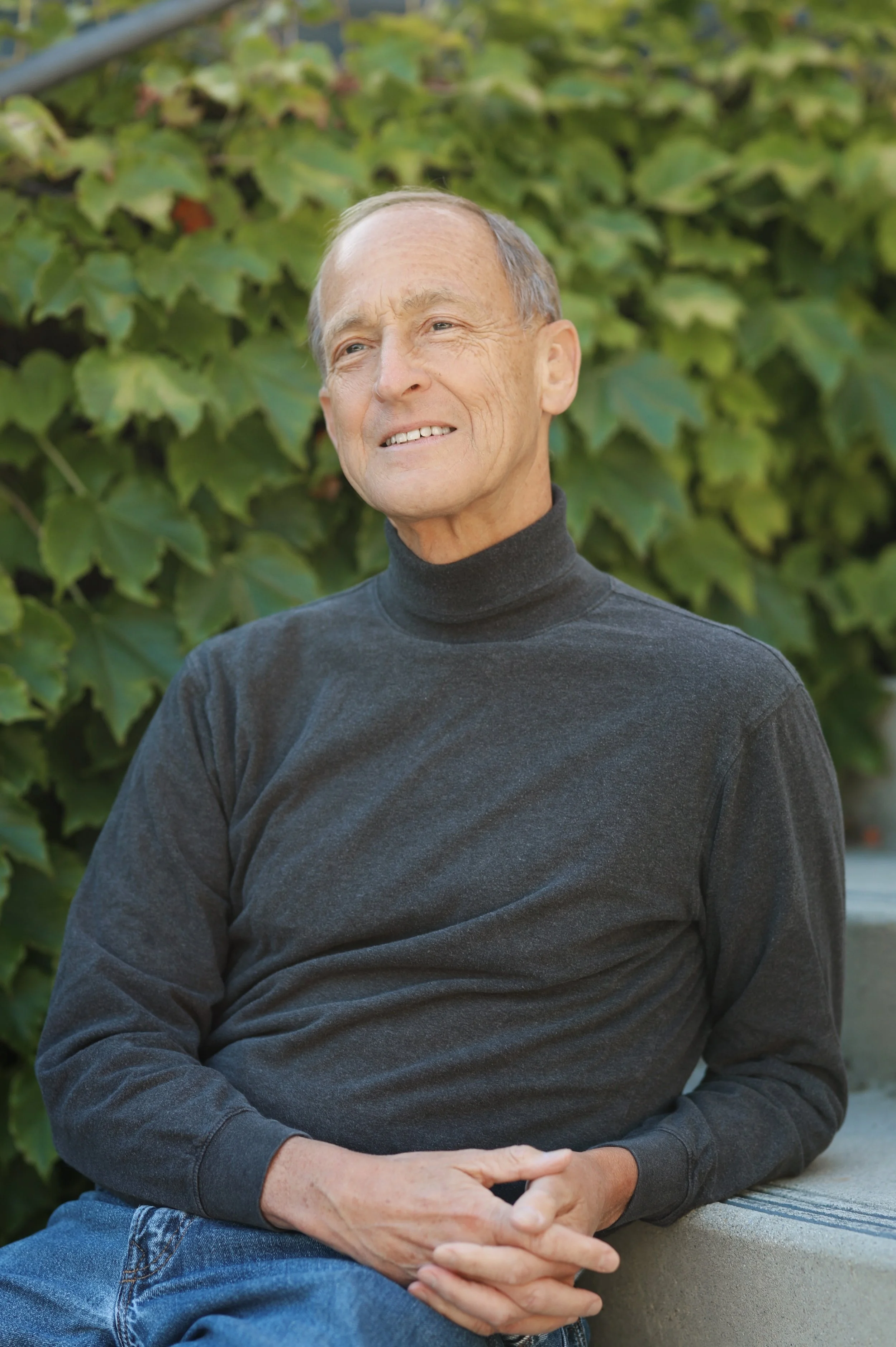 An elderly man with a contemplative expression, seated outdoors in front of green ivy, wearing a black turtleneck and blue jeans.