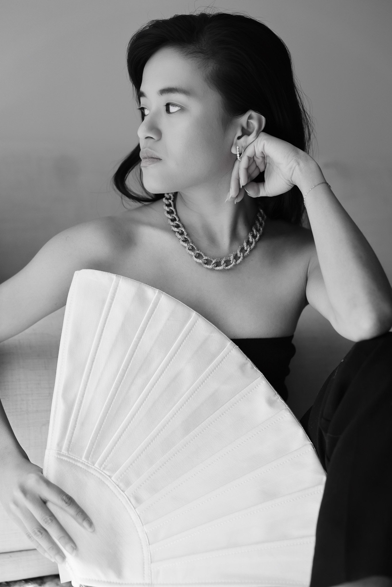 A black and white portrait of a woman with short dark hair, wearing jewelry including a thick chain necklace, earrings, and a thin bracelet. She is holding a large decorative fan partially covering her chest and is looking to the left with a thoughtf