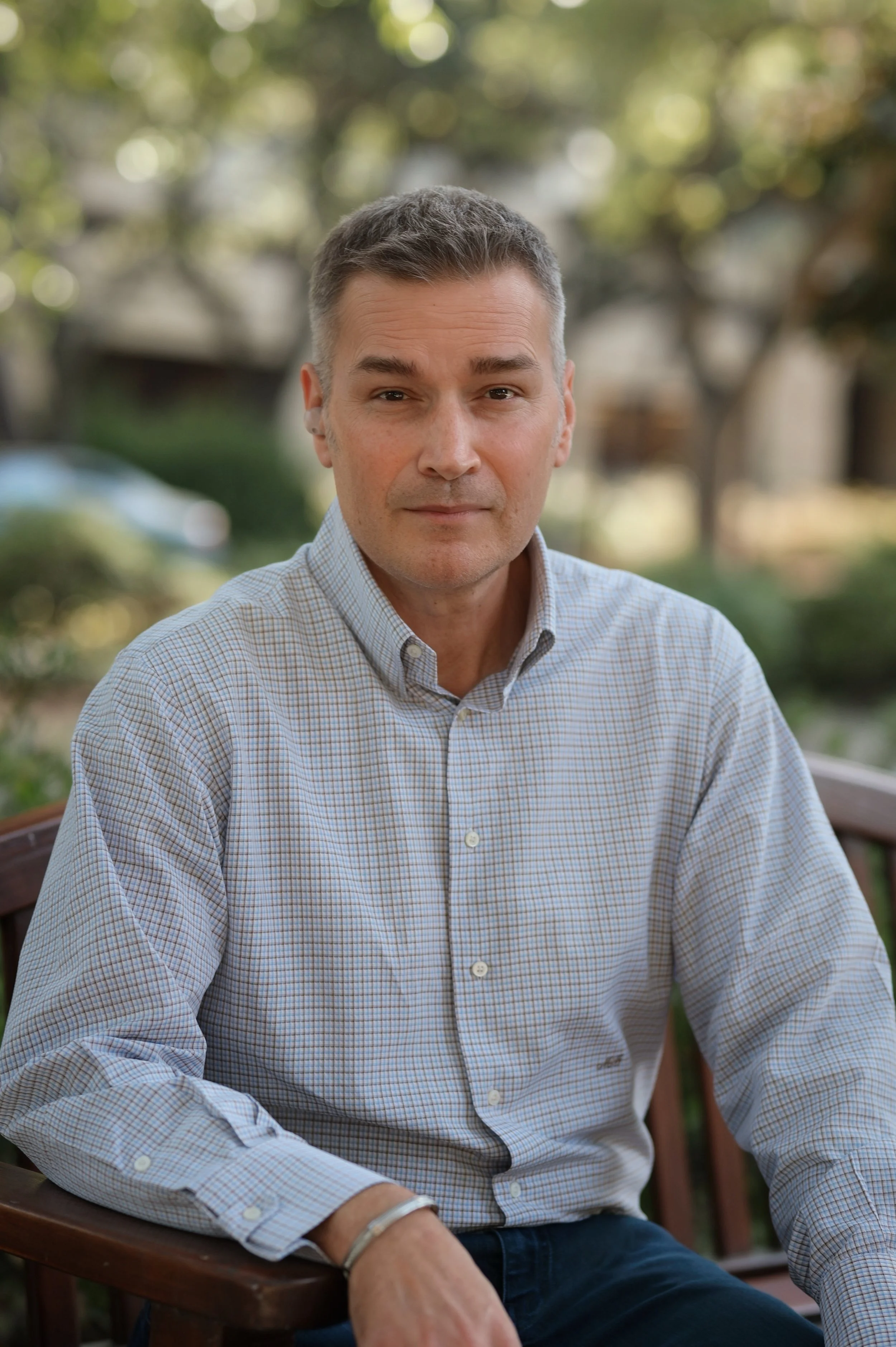 A middle-aged man with short, gray hair sits outdoors on a wooden bench. He wears a light-colored, checkered button-up shirt and is looking directly at the camera with a neutral expression. The background is blurred with trees and greenery.