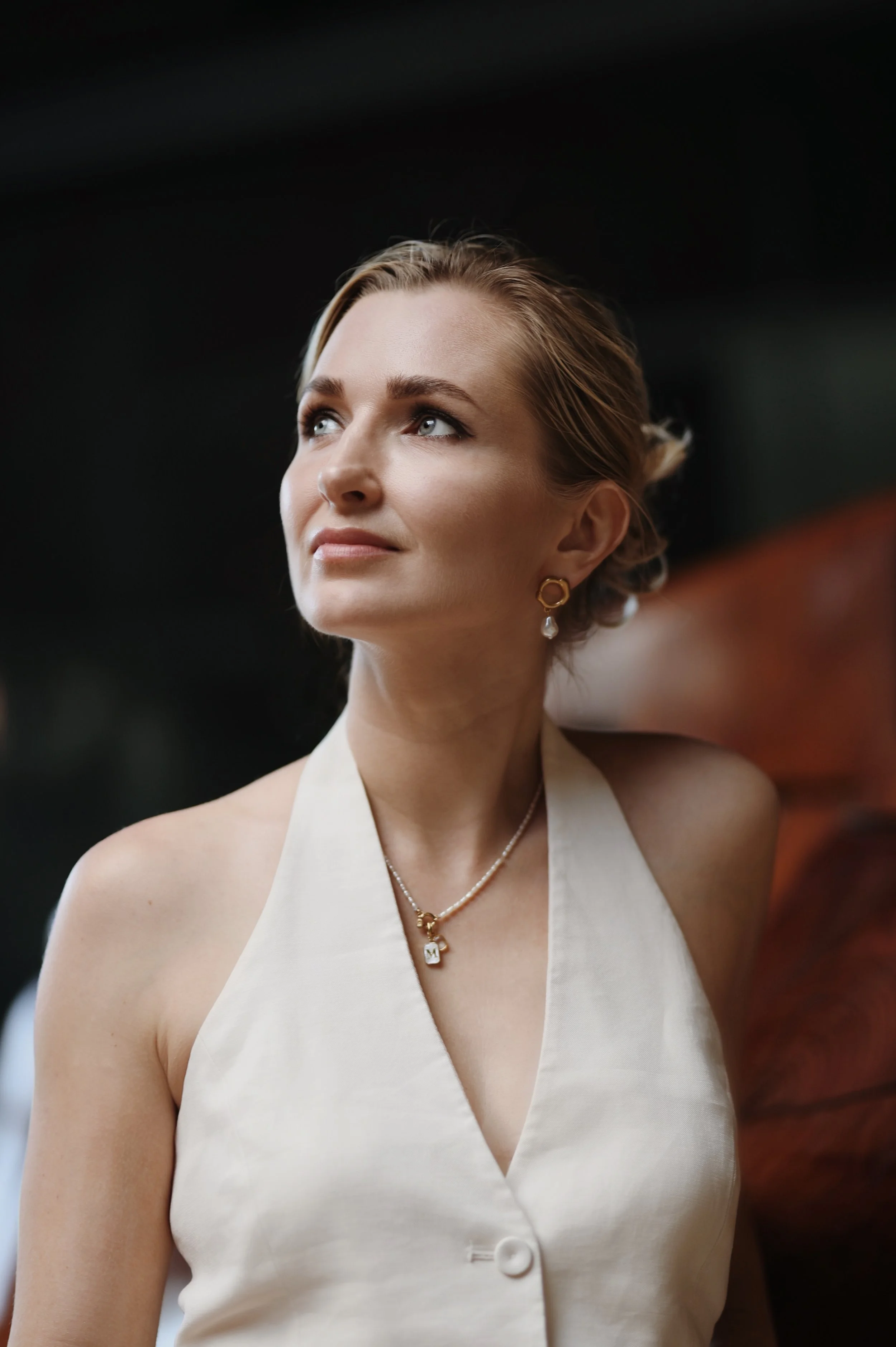 A woman with styled hair wearing elegant jewelry and a sleeveless cream-colored top, looking thoughtfully upward.