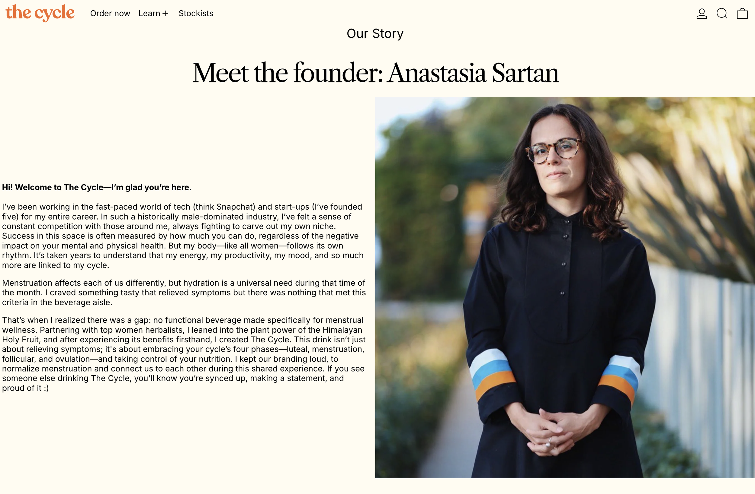 Portrait of a woman with curly dark hair wearing glasses and a dark shirt with colorful striped sleeves, standing outdoors with blurred trees and foliage in the background.