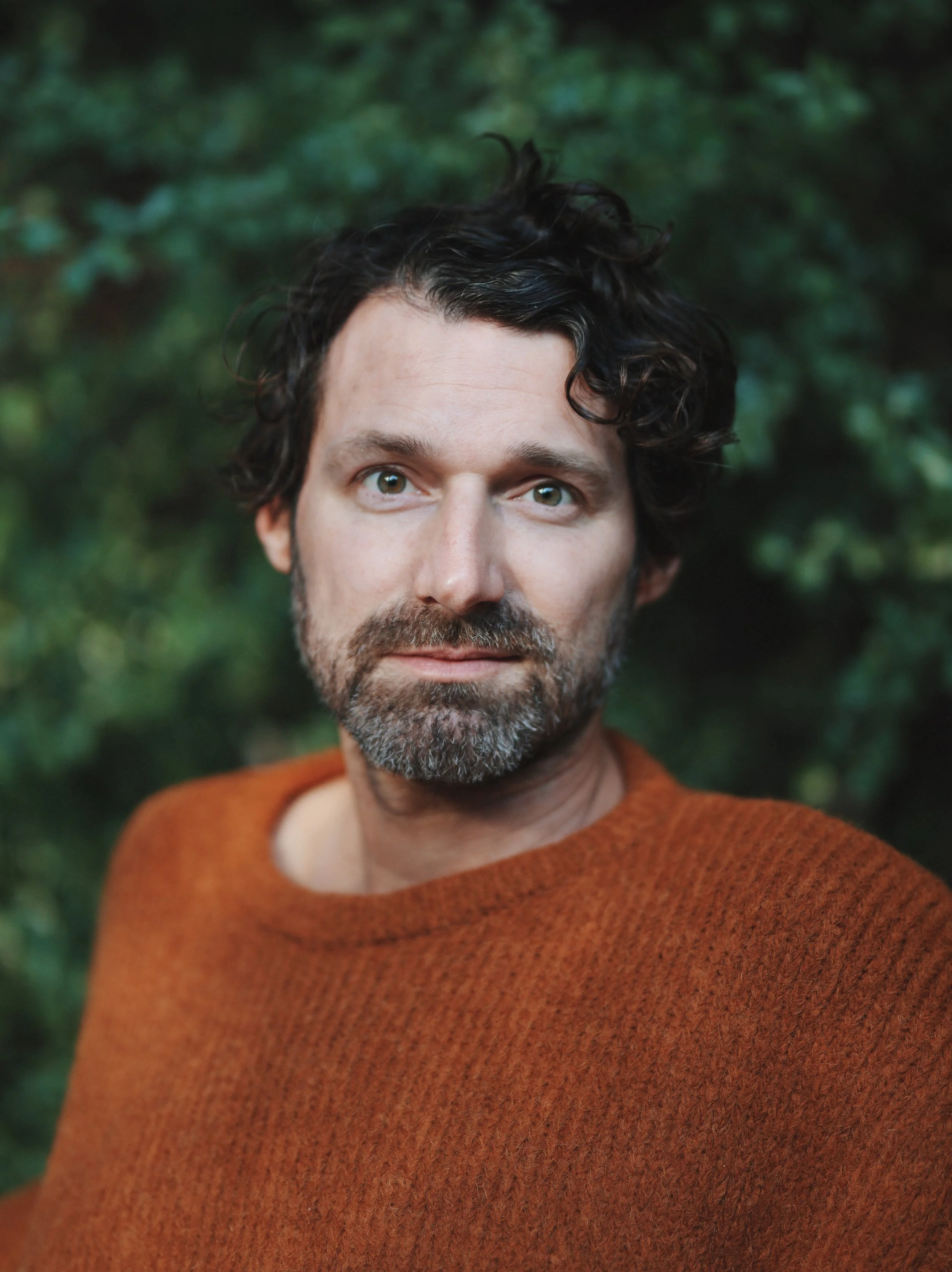 A man with dark, curly hair and a beard, wearing an orange-brown sweater, standing outdoors with blurred green foliage in the background.
