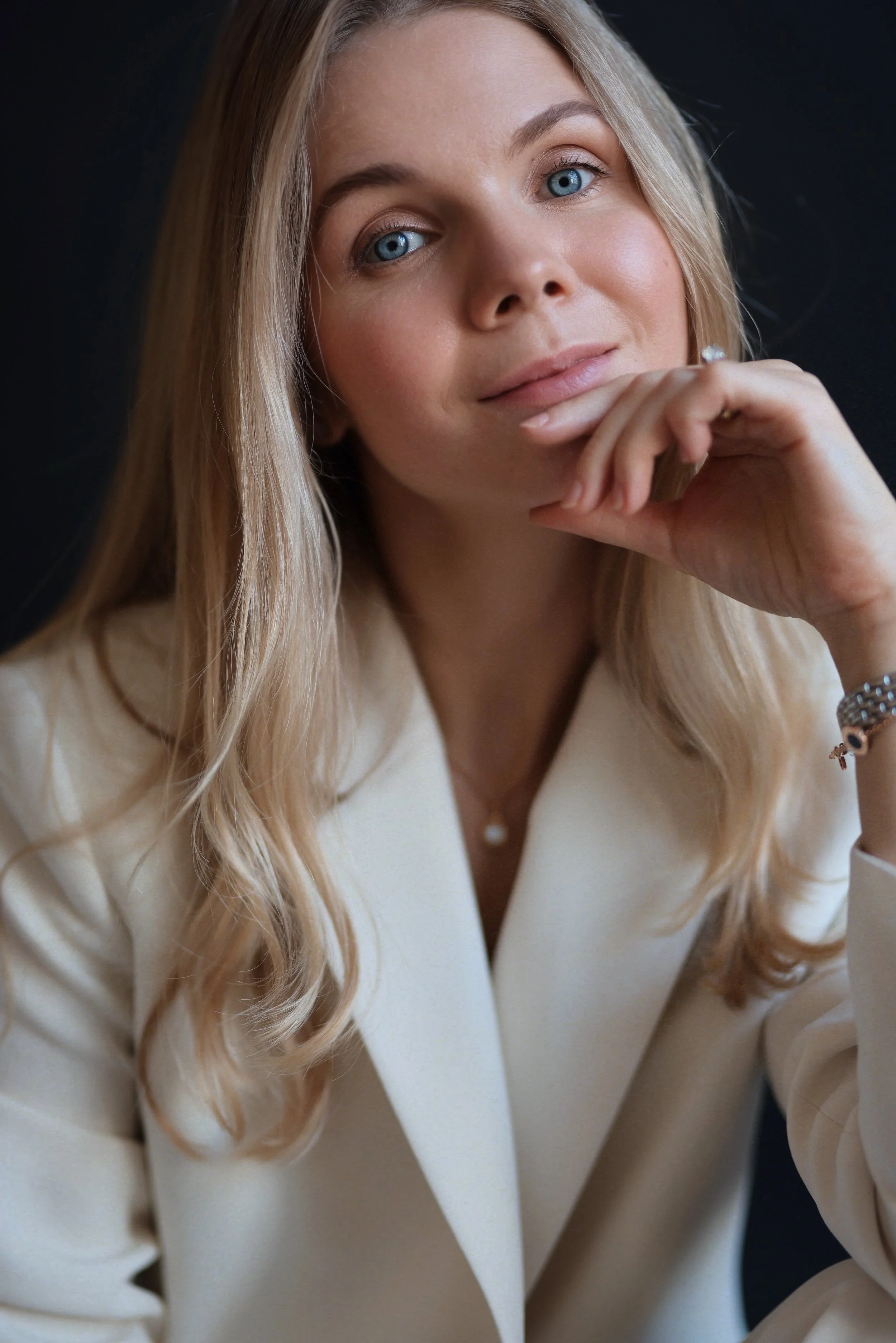 A woman with long blonde hair and blue eyes, wearing a white blazer, resting her chin on her hand, looking into the camera with a slight smile.