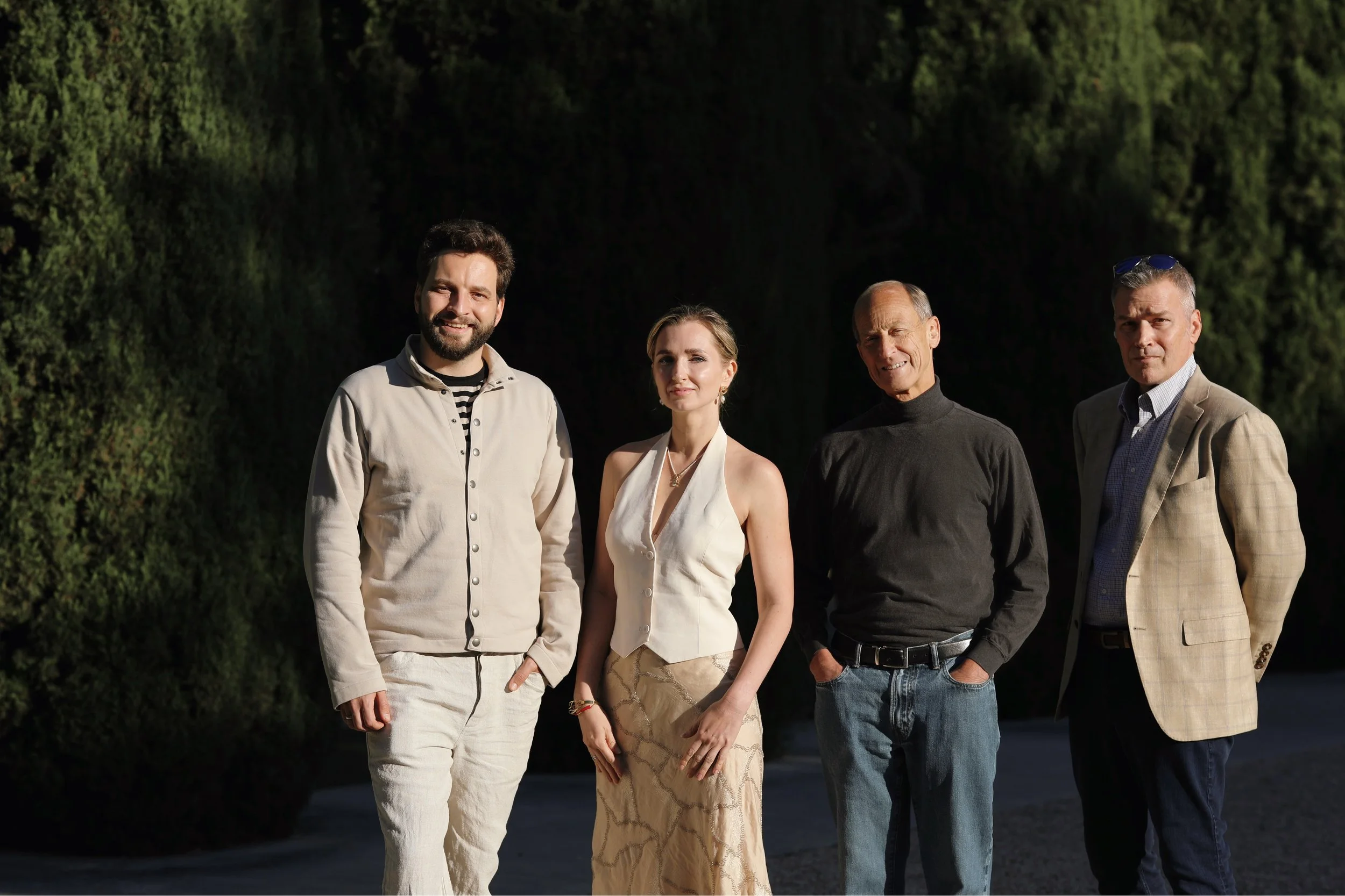 Four people standing outdoors in front of a green wooded background, dressed in casual and semi-formal clothing, smiling at the camera.