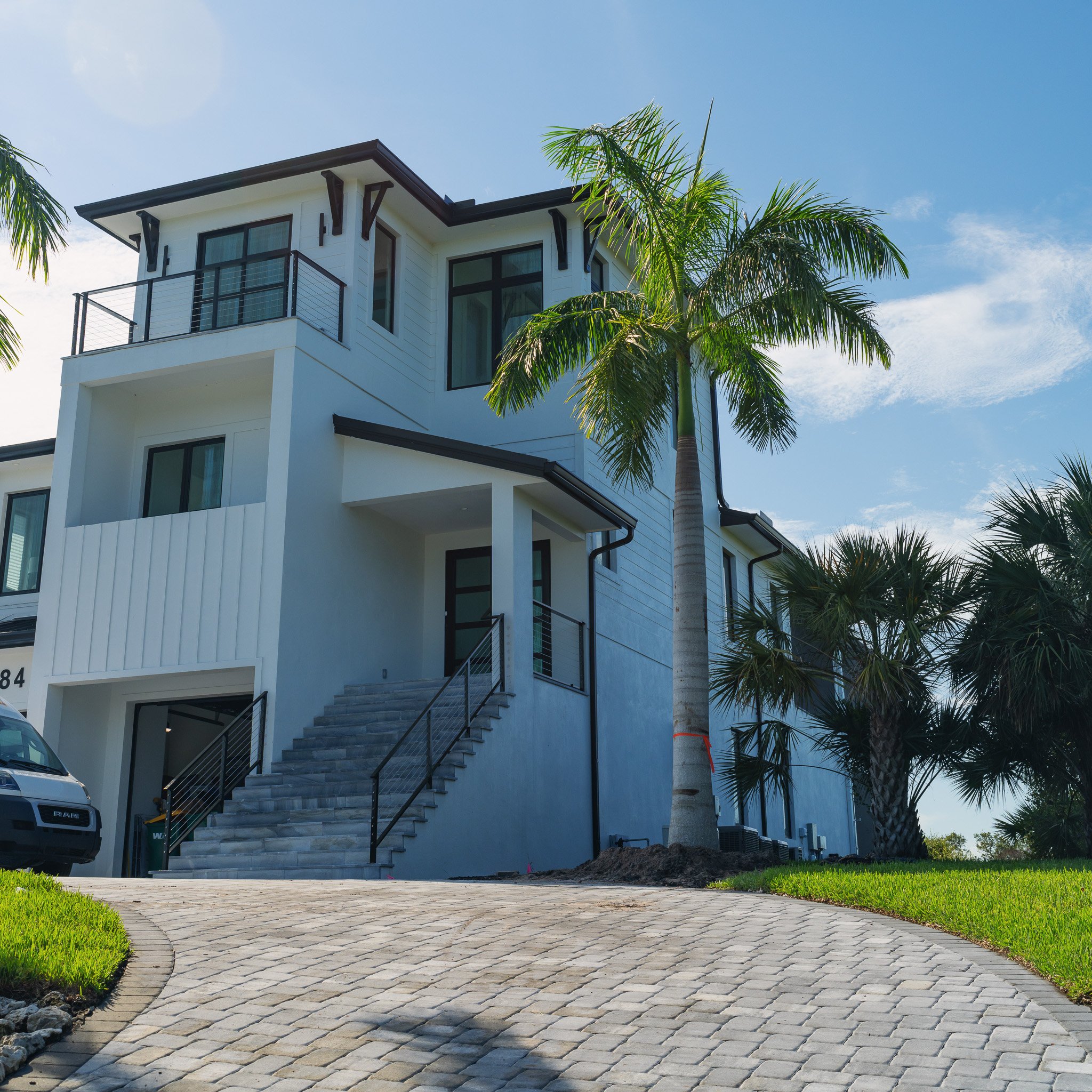 Modern white multi-story house with black accents, palm trees, a curved stone driveway, and a staircase leading to an entrance
