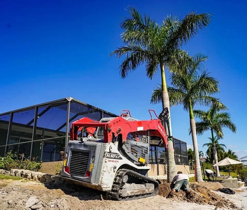 A construction site with a red and white compact excavator parked on dirt, surrounded by palm trees and a modern glass building in the background.