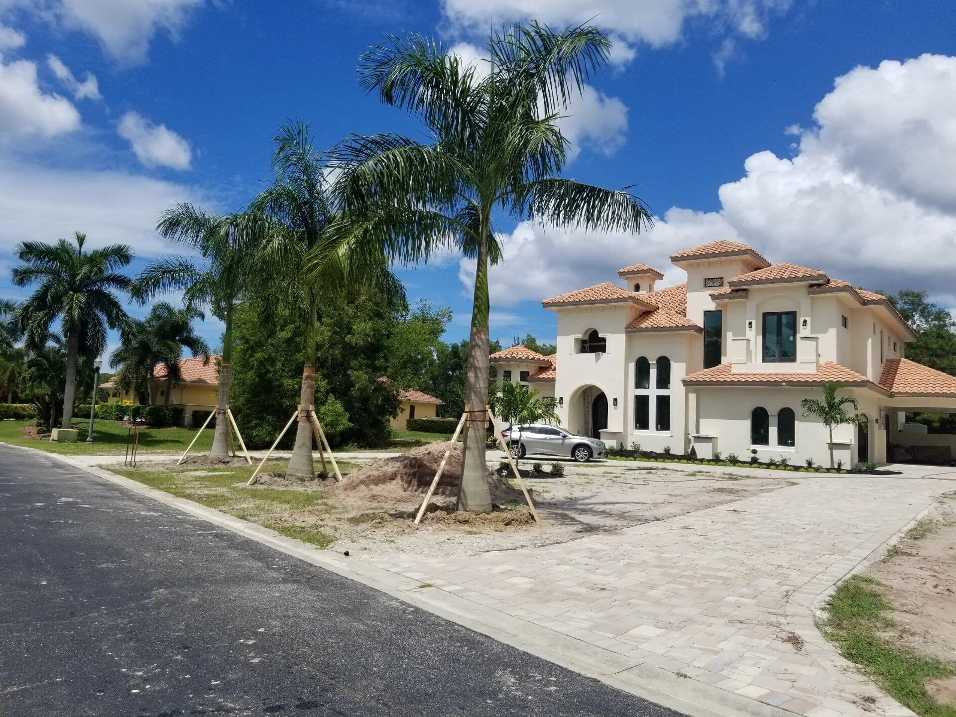 A large white Mediterranean-style house with red tile roof, surrounded by palm trees, under a partly cloudy blue sky.