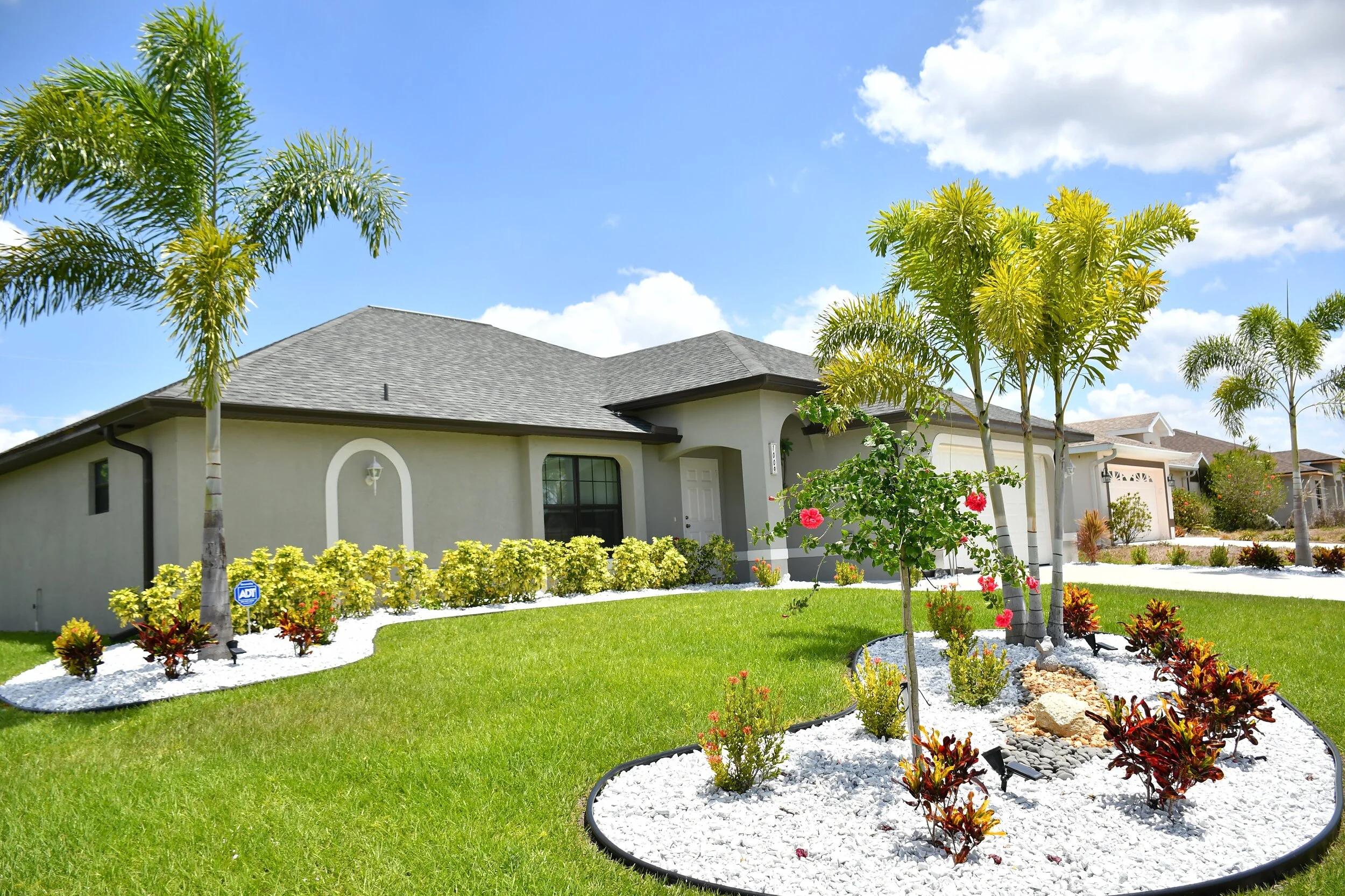 Front yard of a suburban house during the day with green lawn, palm trees, flowering bushes, and a white gravel landscape.