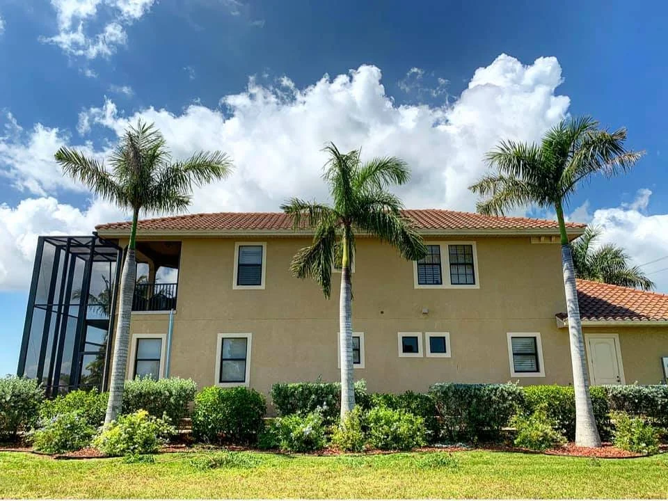 A two-story beige house with a red tile roof, surrounded by palm trees and green shrubbery, under a partly cloudy blue sky.