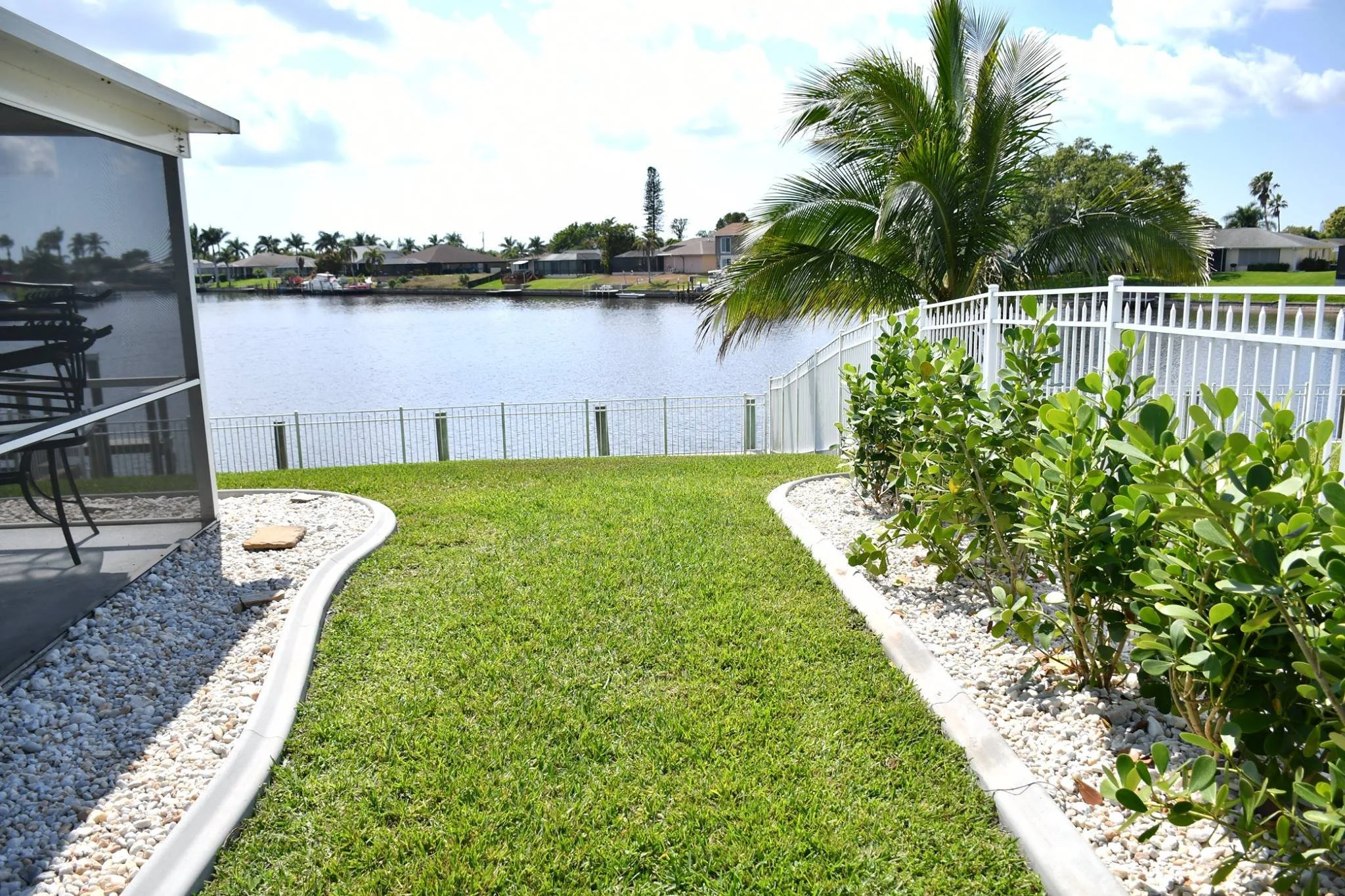View of a backyard with green grass, landscaped bushes with pebbles along the edges, a white fence, a palm tree, and a water body with houses on the other side under a partly cloudy sky.