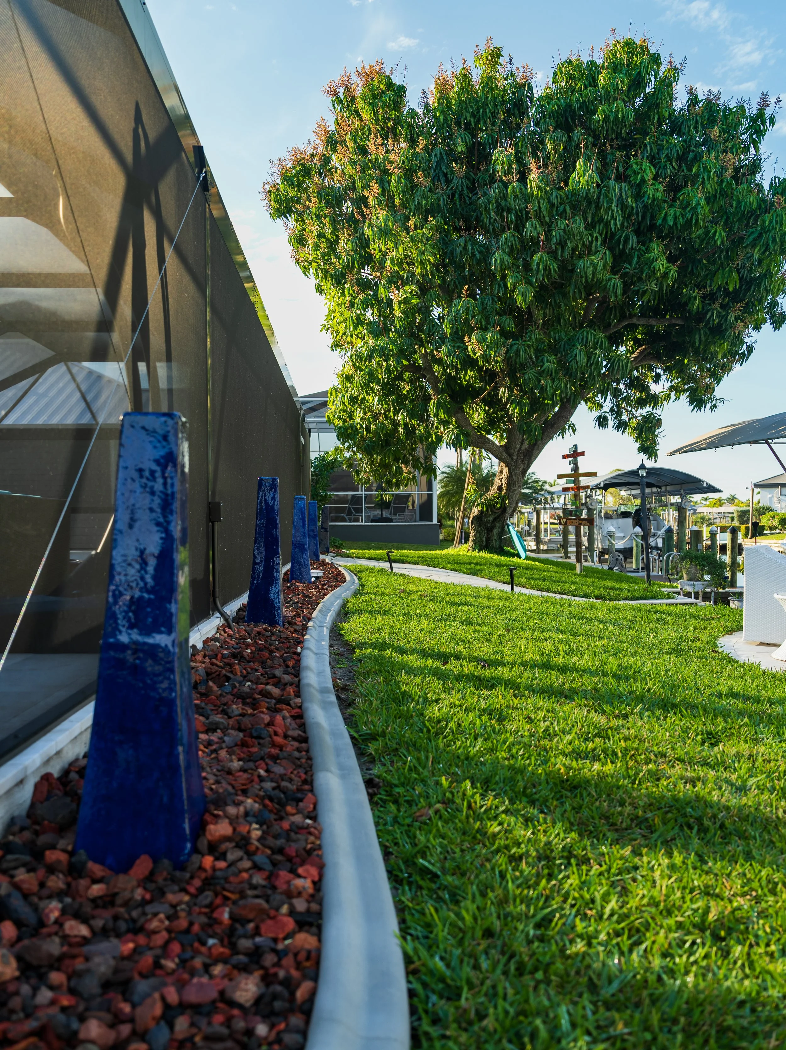 A landscaped outdoor area with a large tree, a curved lawn, and modern outdoor furniture. Blue decorative stones are lined along a building wall, and a signpost is visible near the tree.