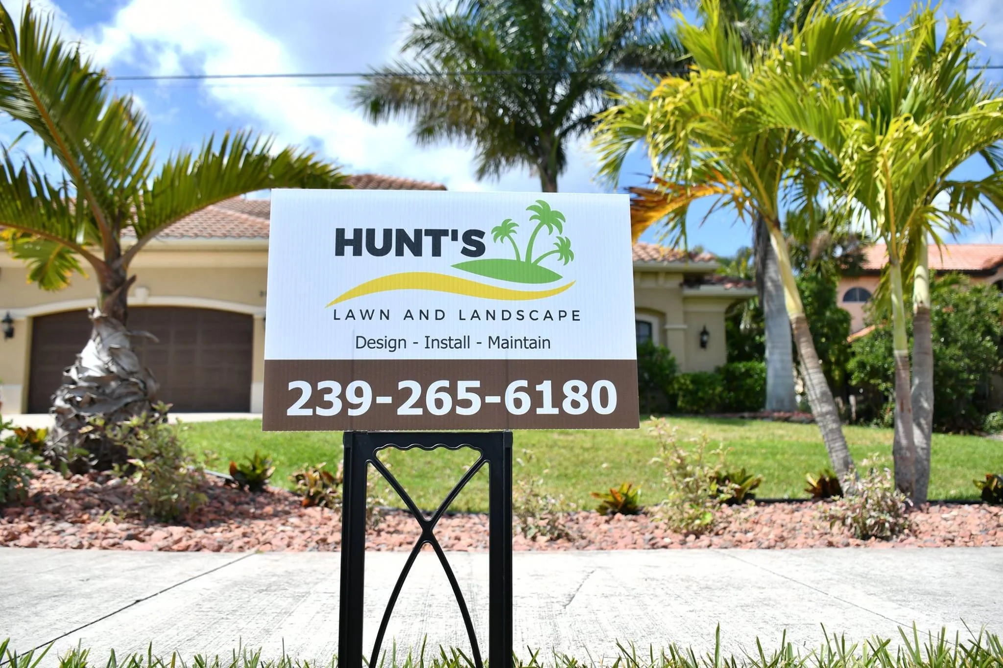 Sign for Hunt's Lawn and Landscape in front yard with palm trees and a house in the background.