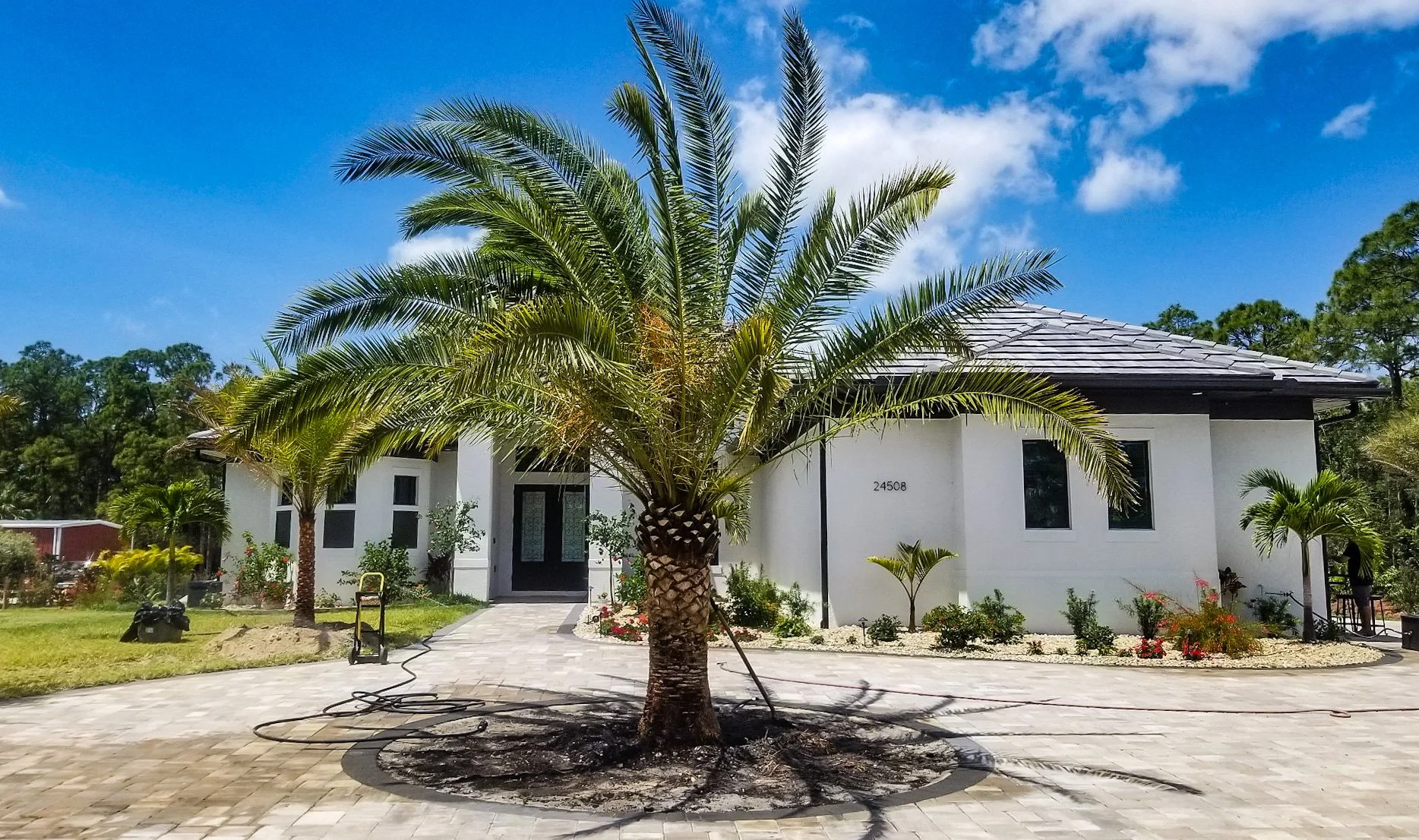 Front view of a modern white house with palm trees in the yard, a paved driveway, and a blue sky with clouds.