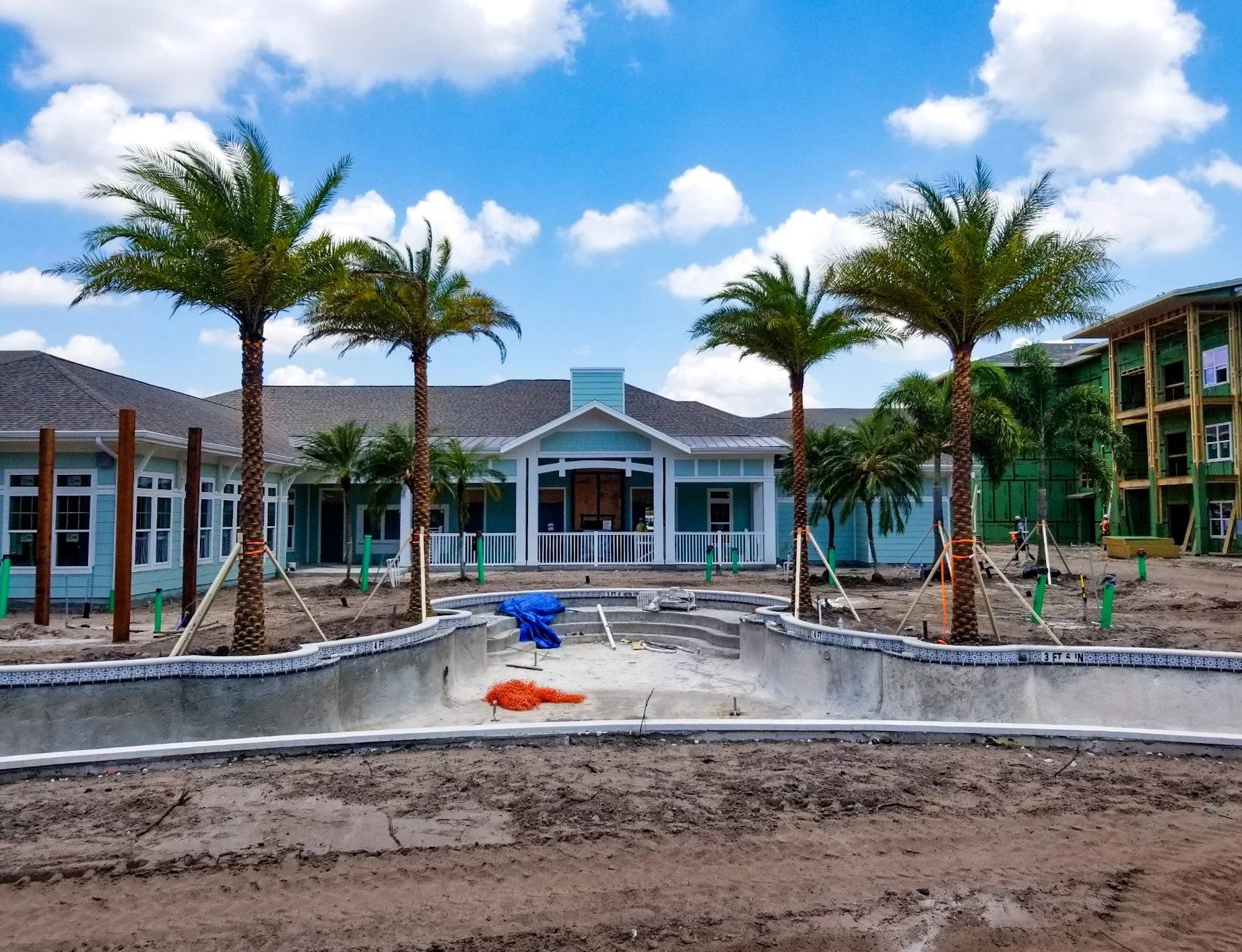 Construction site of a residential swimming pool with tall palm trees, a sunny sky, and a partly built house in the background.