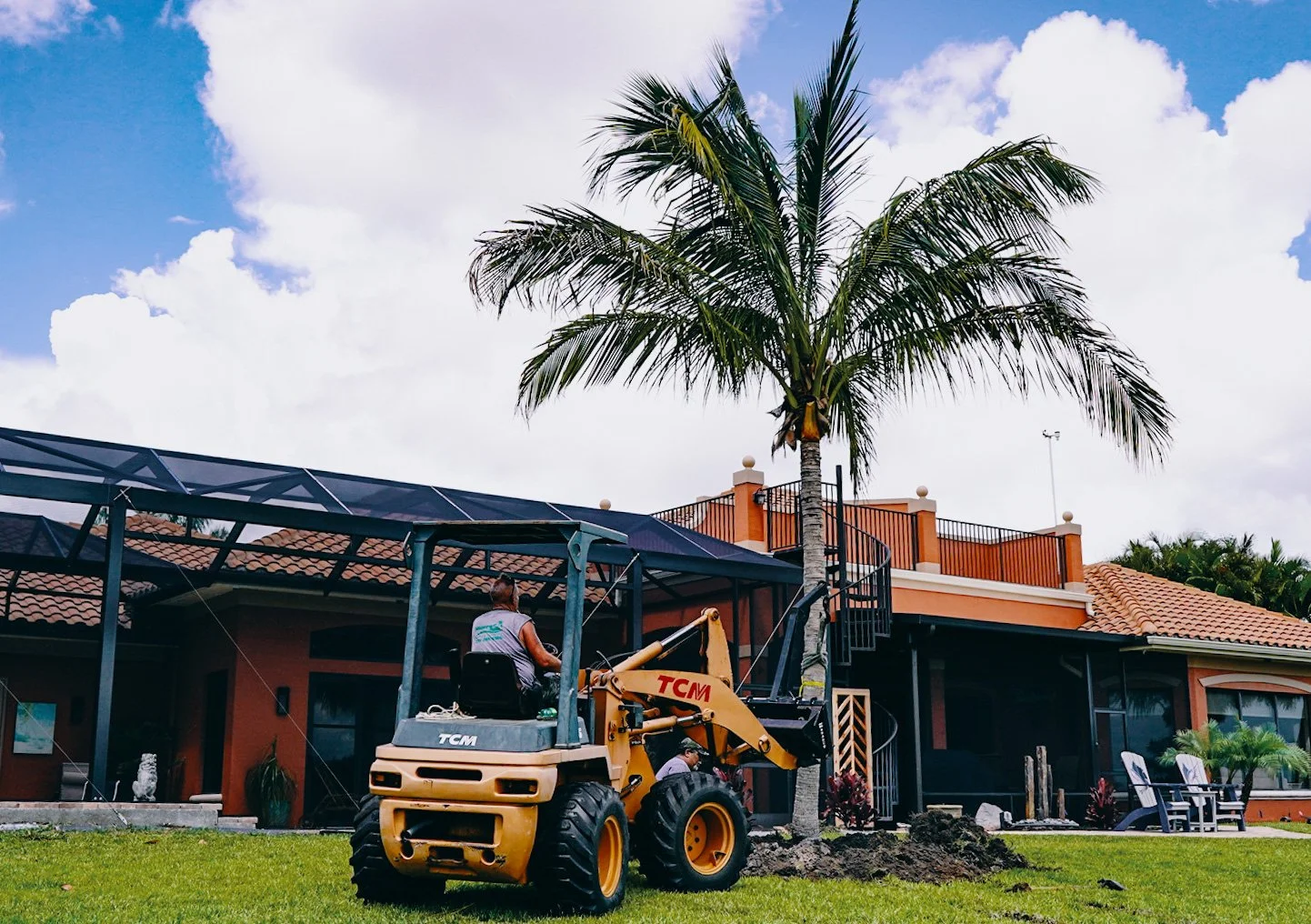 Construction worker operating a small excavator on a lawn in front of a house with a large palm tree and a roof covered with solar panels.