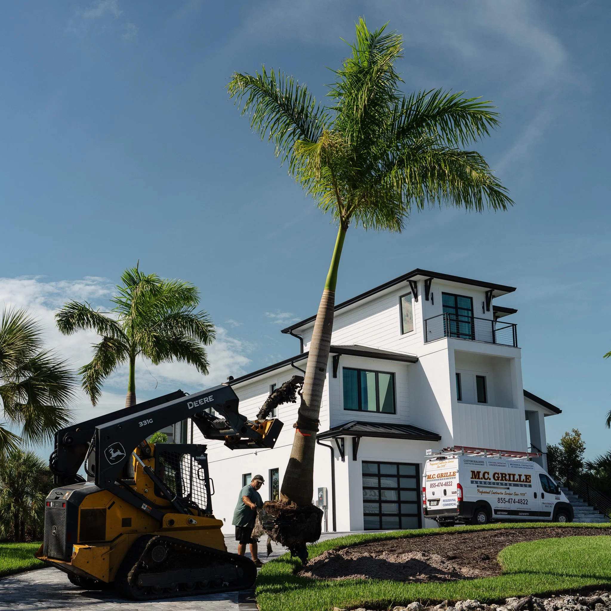 A worker using equipment to transplant a large palm tree in front of a modern white house with multiple stories, black accents, and large windows. A white work van with red lettering is parked nearby.