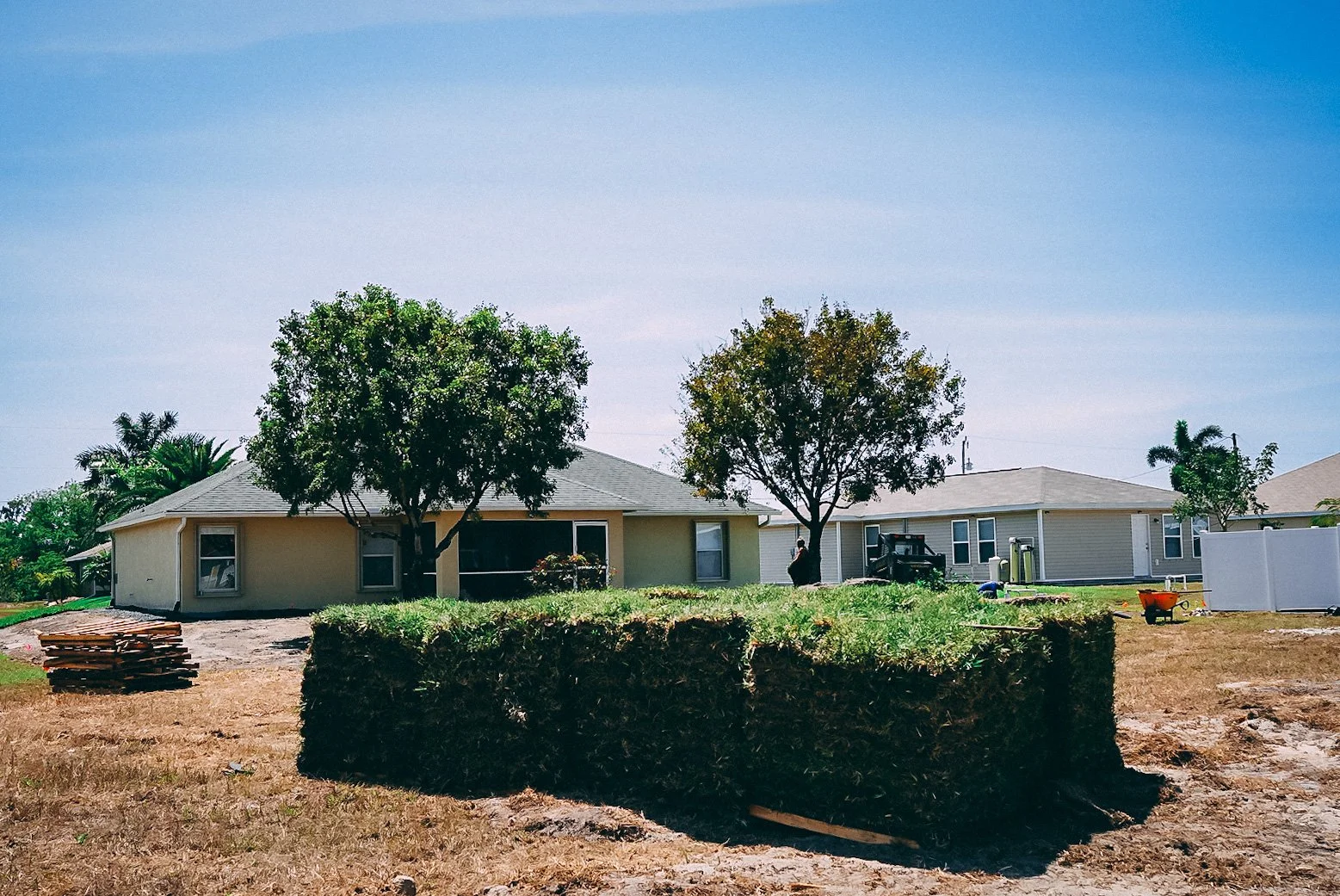 A backyard with two single-story houses, trees, and a large newly dug hole with grass on top in the foreground, construction materials, and tools scattered around.