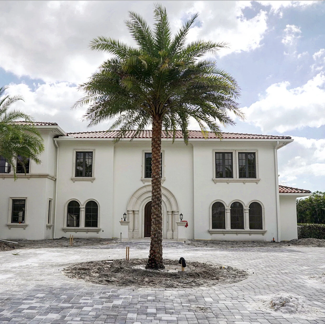 A large house with a white facade and arched windows, with a tall palm tree in front, under a partly cloudy sky.