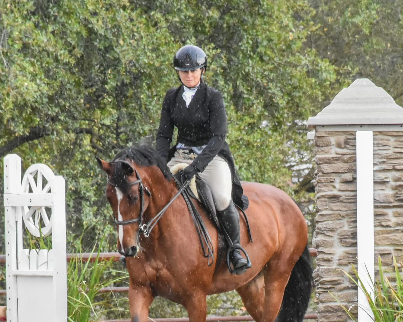 A woman riding a horse during a rainstorm near a stone and white wooden fence, with trees in the background.