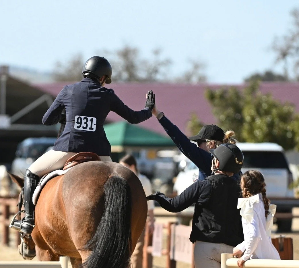 A person on horseback wearing a black helmet and riding jacket high-fives a woman in a black cap and jacket, with children nearby, at an outdoor event.