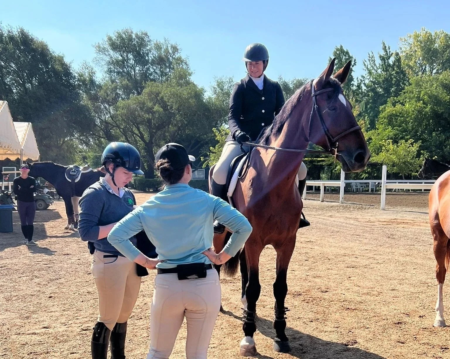 A young woman in riding attire sitting on a horse, talking to two women in riding gear and helmets, with other people and horses in the background at an outdoor riding arena.