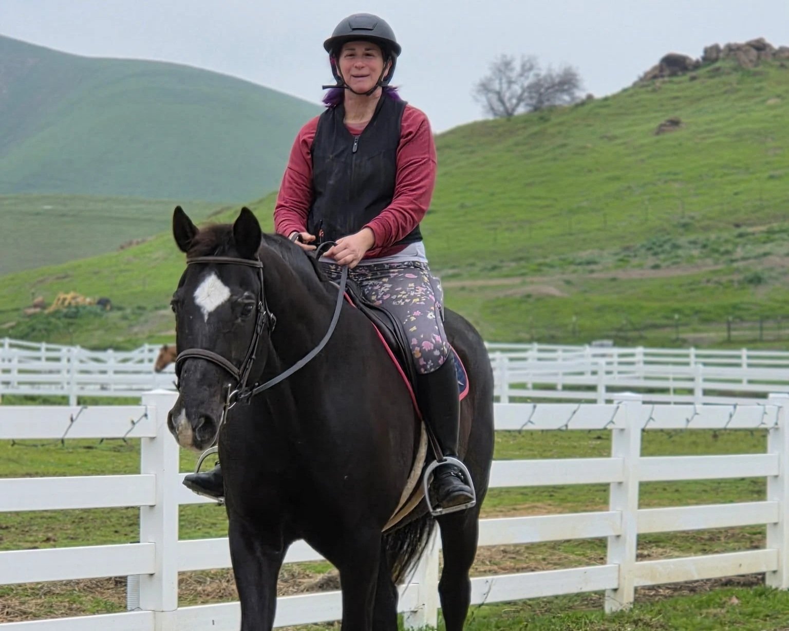 A woman riding a black horse along a white fence with green hills in the background.